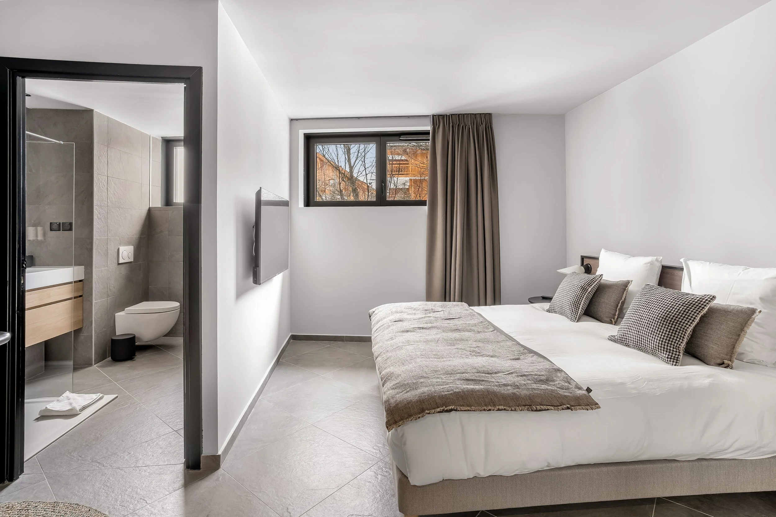 A modern hotel room with a bed, pillows, a window with beige curtains, and an adjacent bathroom visible through an open door.
