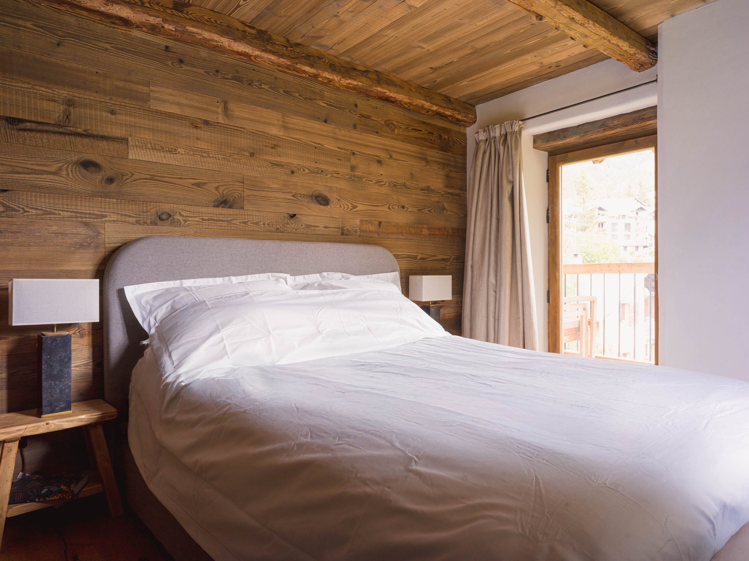 Une chambre à coucher avec un lit recouvert de draps blancs, une tête de lit beige, deux petites tables de chevet avec des lampes, des murs et un plafond lambrissés, une fenêtre aux rideaux beiges, et une vue sur un balcon et des arbres à l'extérieur.