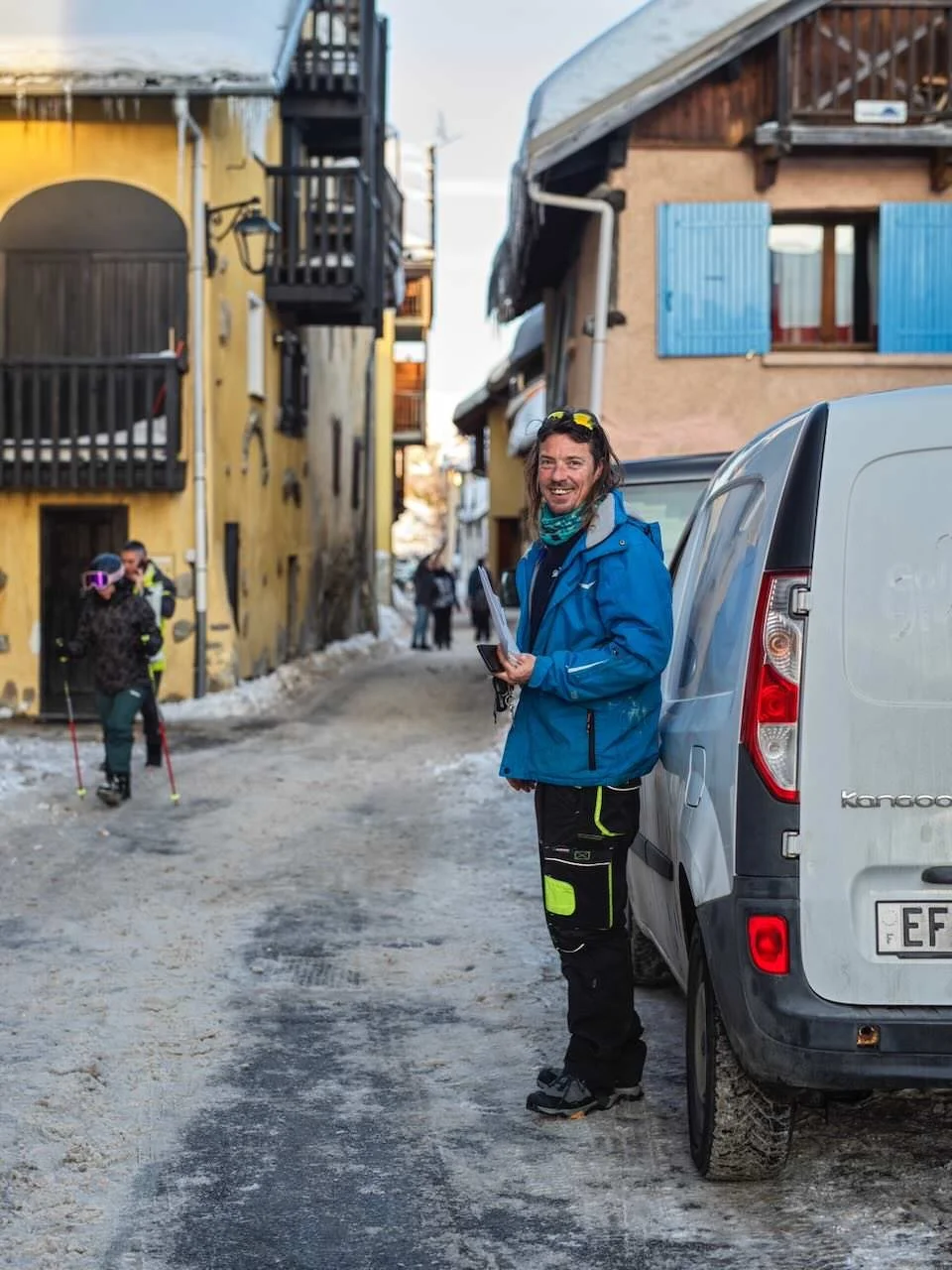 A smiling man wearing outdoor gear, standing beside a gray van on a snowy, narrow street with colorful buildings in the background. People are seen walking and skiing in the distance.