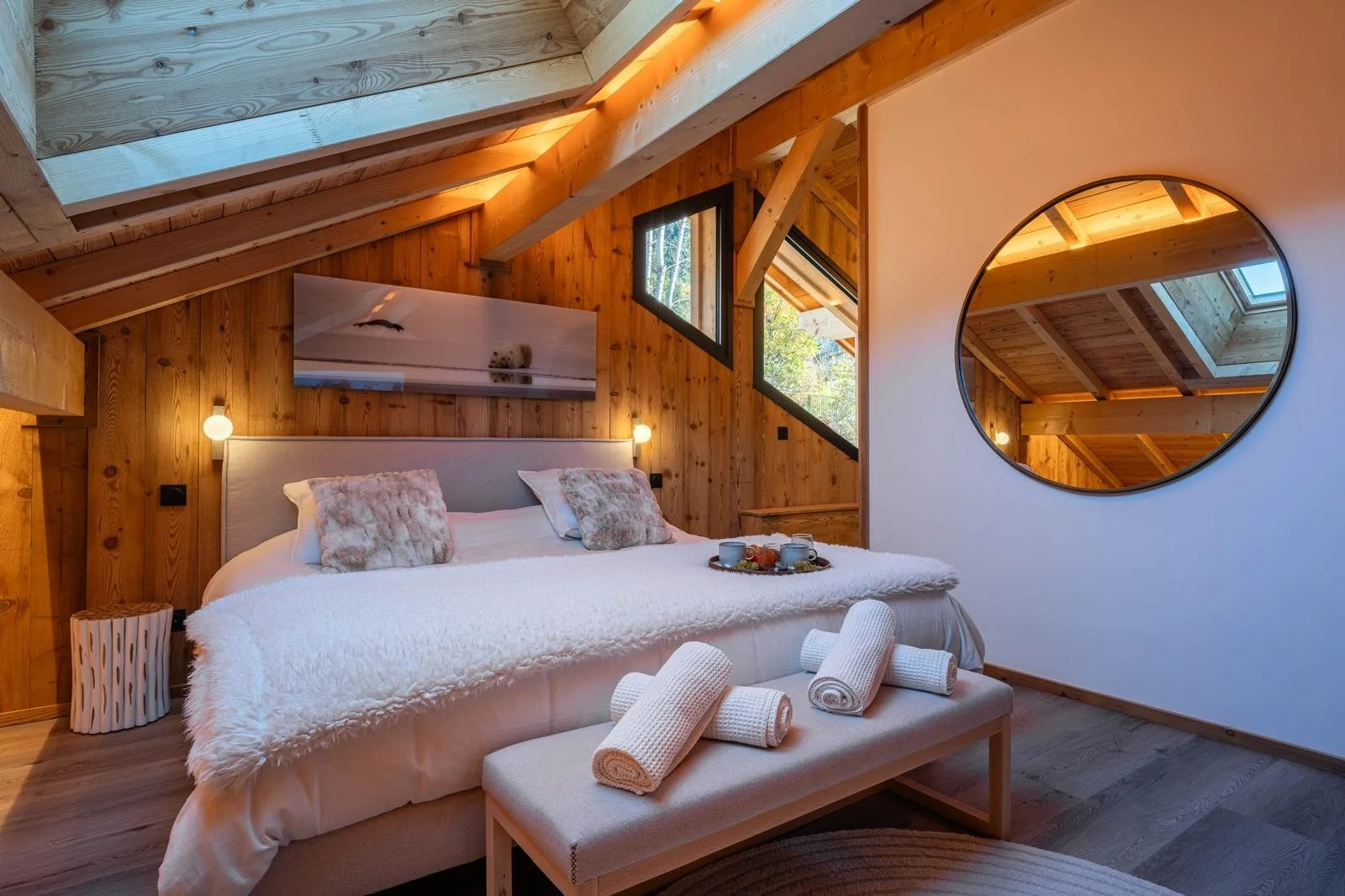 Cozy bedroom in a wooden cabin with a large bed, cream-colored bedspread, decorative pillows, and a tray with breakfast. The room has sloped ceilings, several windows, warm lighting, and a round mirror on the wall.