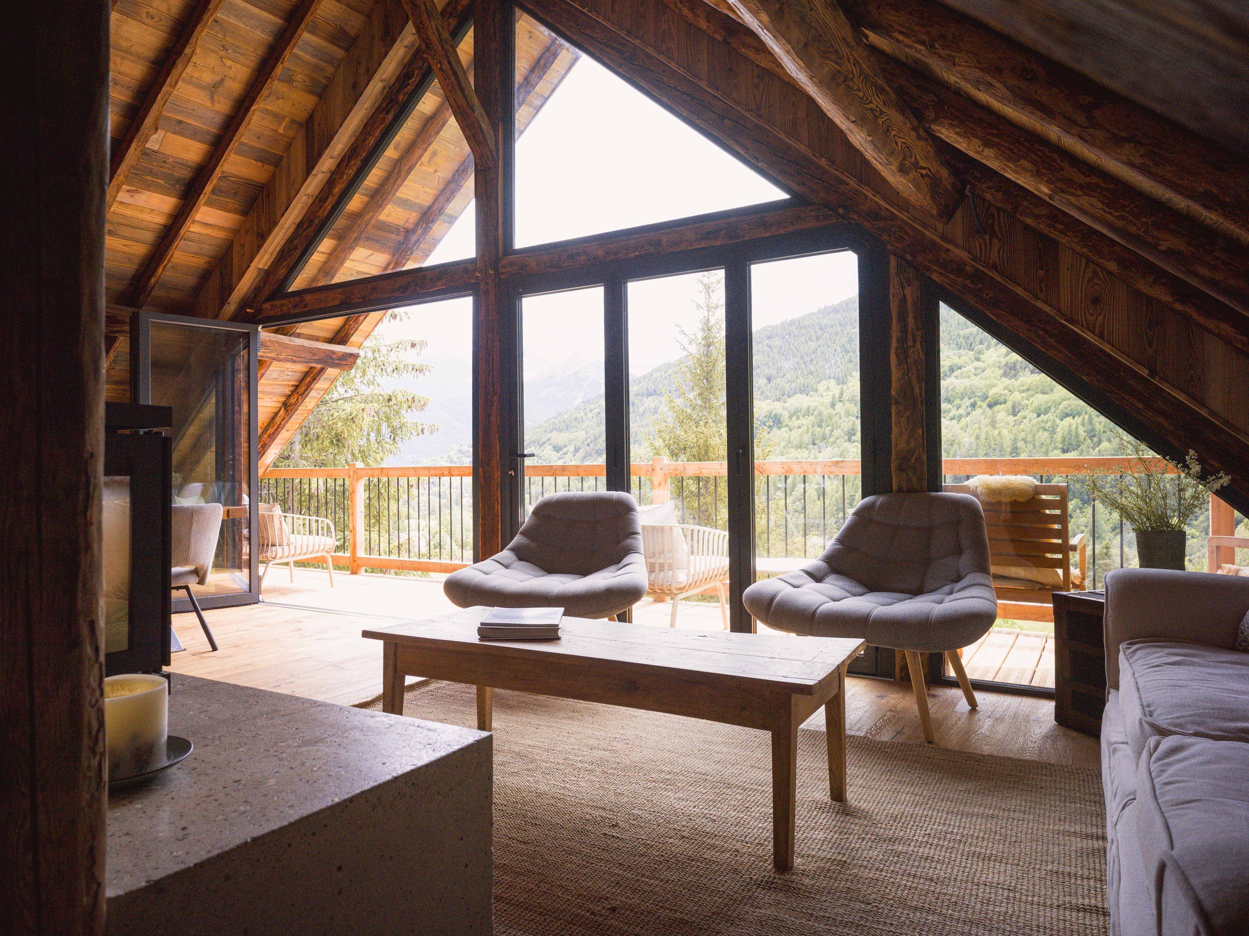 Un coin salon doté de grandes baies vitrées offrant une vue sur les montagnes, avec des poutres apparentes, un tapis beige, deux fauteuils gris, une table basse en bois sur laquelle sont posés des magazines, le tout dans une ambiance chaleureuse.