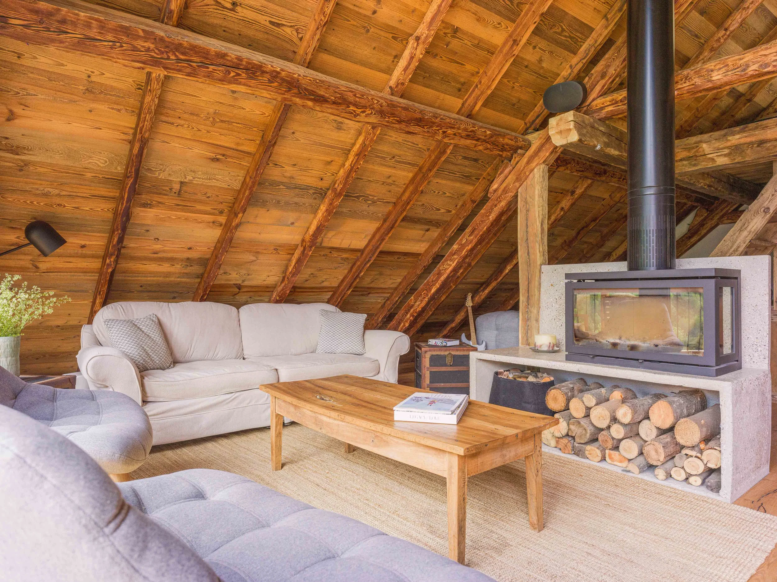 Salon avec un plafond rustique en bois, un canapé blanc orné de coussins, une table basse en bois sur laquelle sont posés des livres, un poêle à bois avec des bûches à ses pieds, ainsi qu'une applique murale noire en forme de bougie.