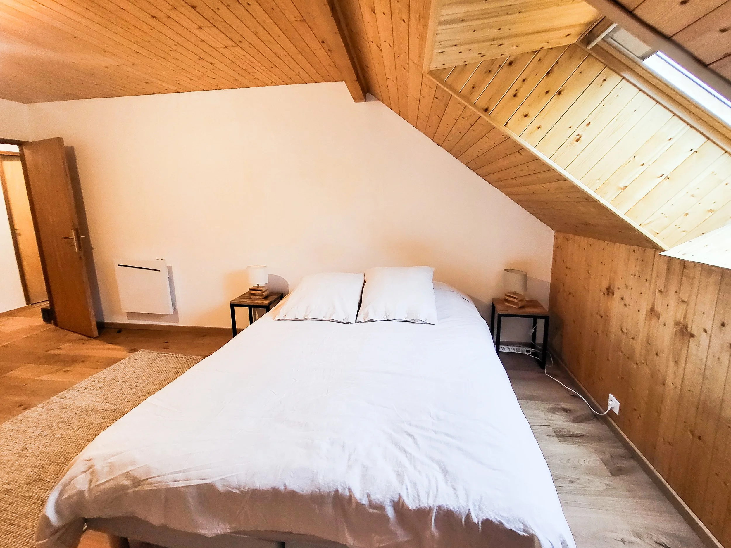 A bedroom with a bed, white bedding, and two pillows, flanked by small black nightstands with lamps, under a sloped wooden ceiling with a skylight.