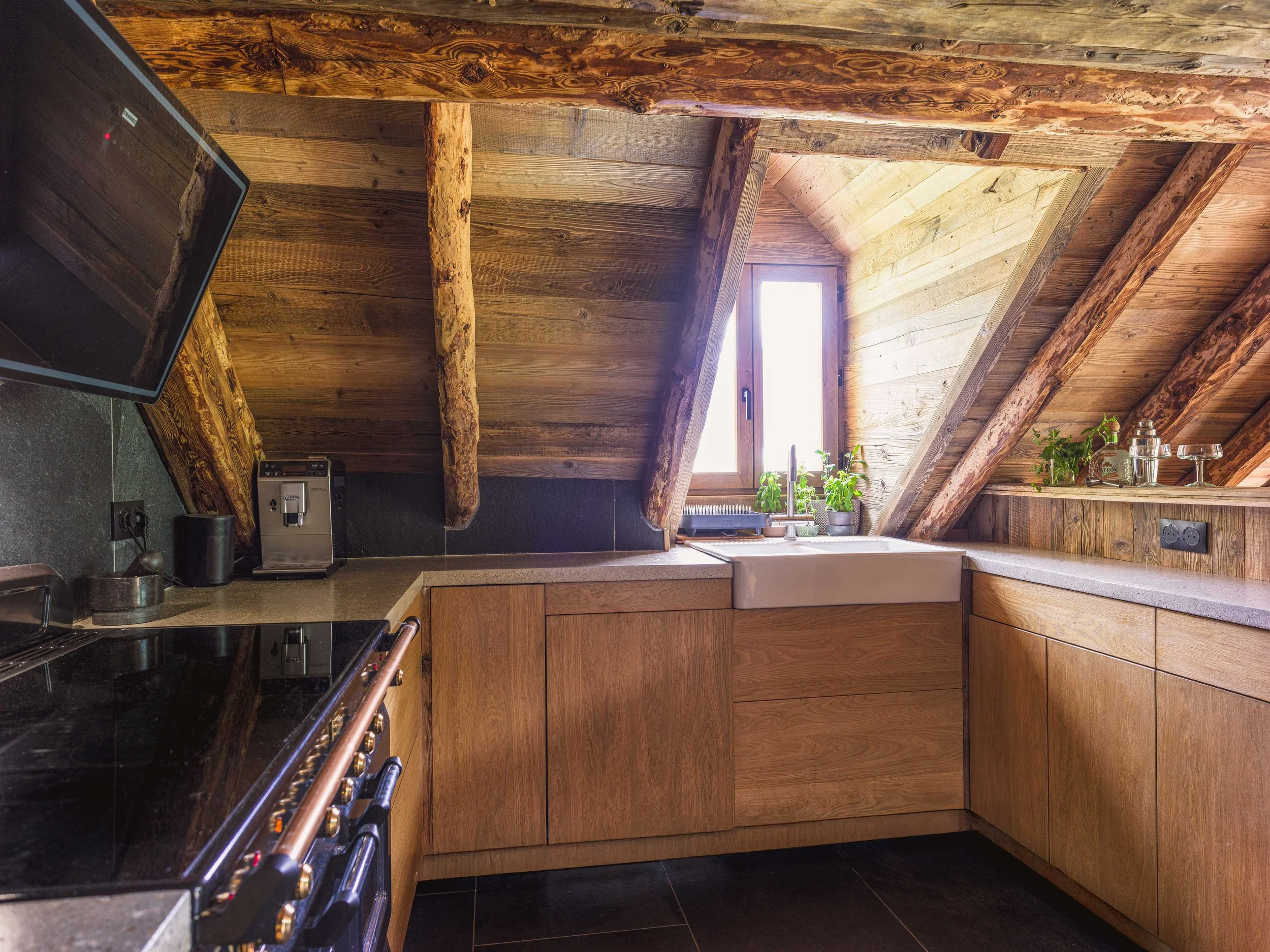 Une cuisine rustique aux murs, au plafond et aux placards en bois, dotée d'un plafond en pente avec une lucarne, d'un plan de travail noir surmonté d'une cuisinière et d'une petite plante sur le rebord de la fenêtre.