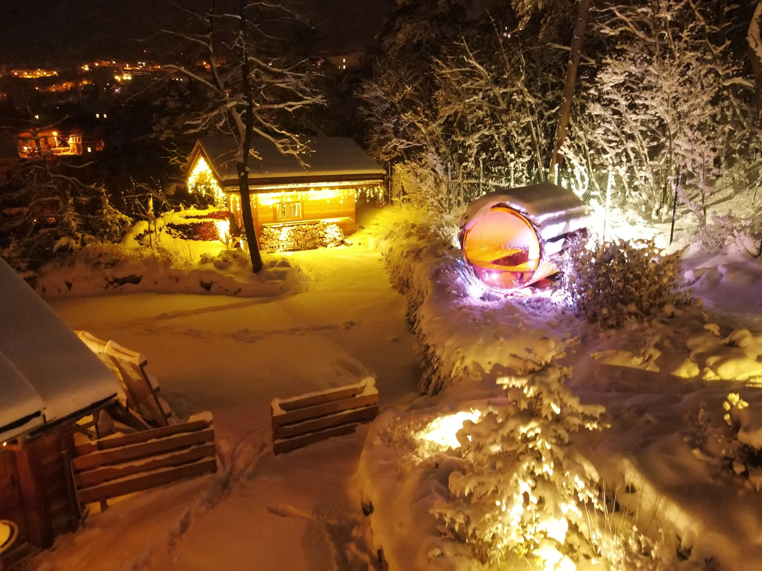 Snow-covered outdoor scene at night with a small lit cabin, illuminated bushes, a glowing enclosed chair, and illuminated snow-covered trees.