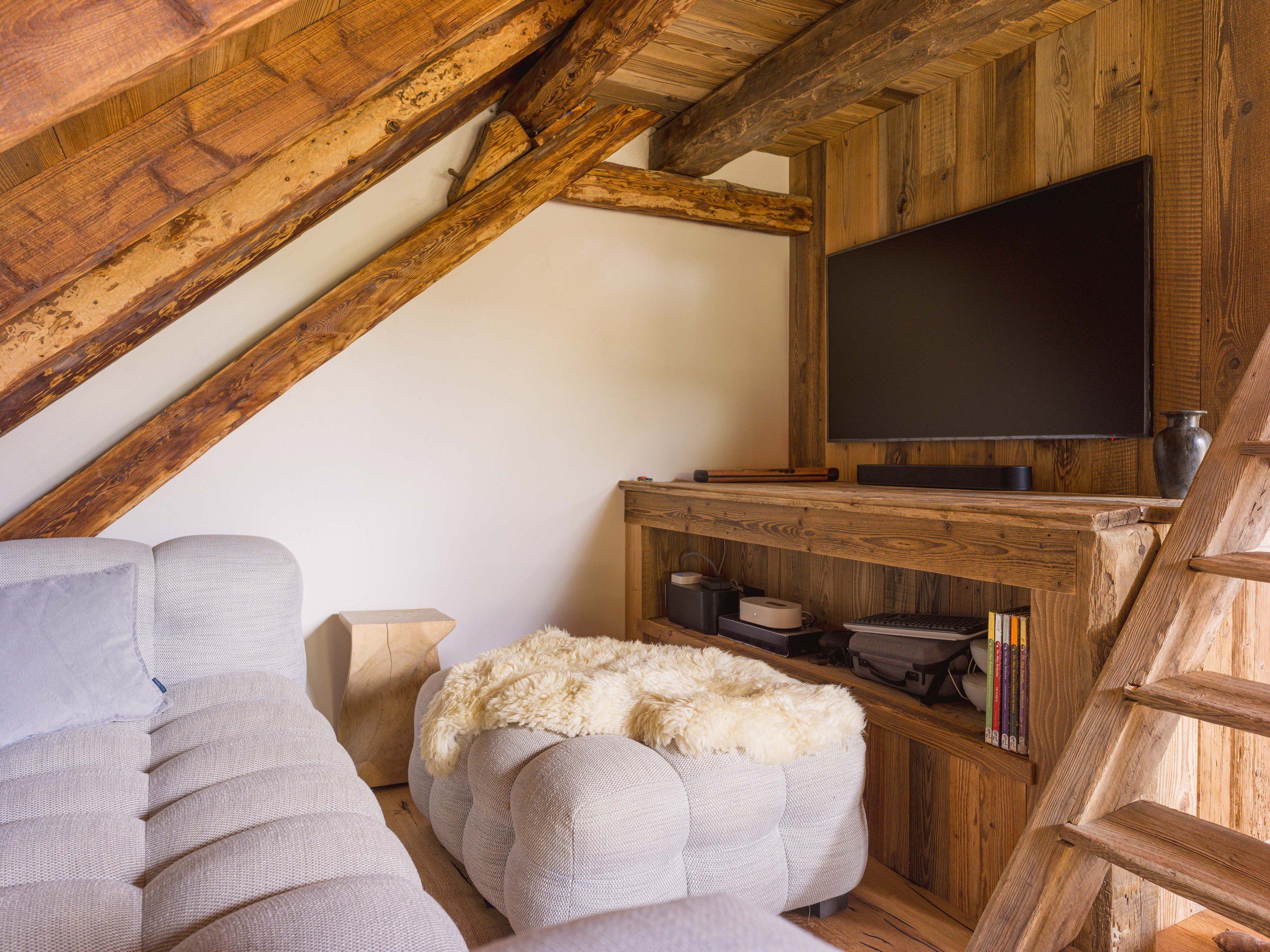 Un coin salon chaleureux avec un canapé modulable capitonné gris, une petite table d'appoint en bois, un plaid moelleux couleur crème, une télévision à écran plat fixée sur un mur lambrissé, une barre de son noire et une collection de livres sur une étagère. La pièce est dotée de poutres apparentes en bois