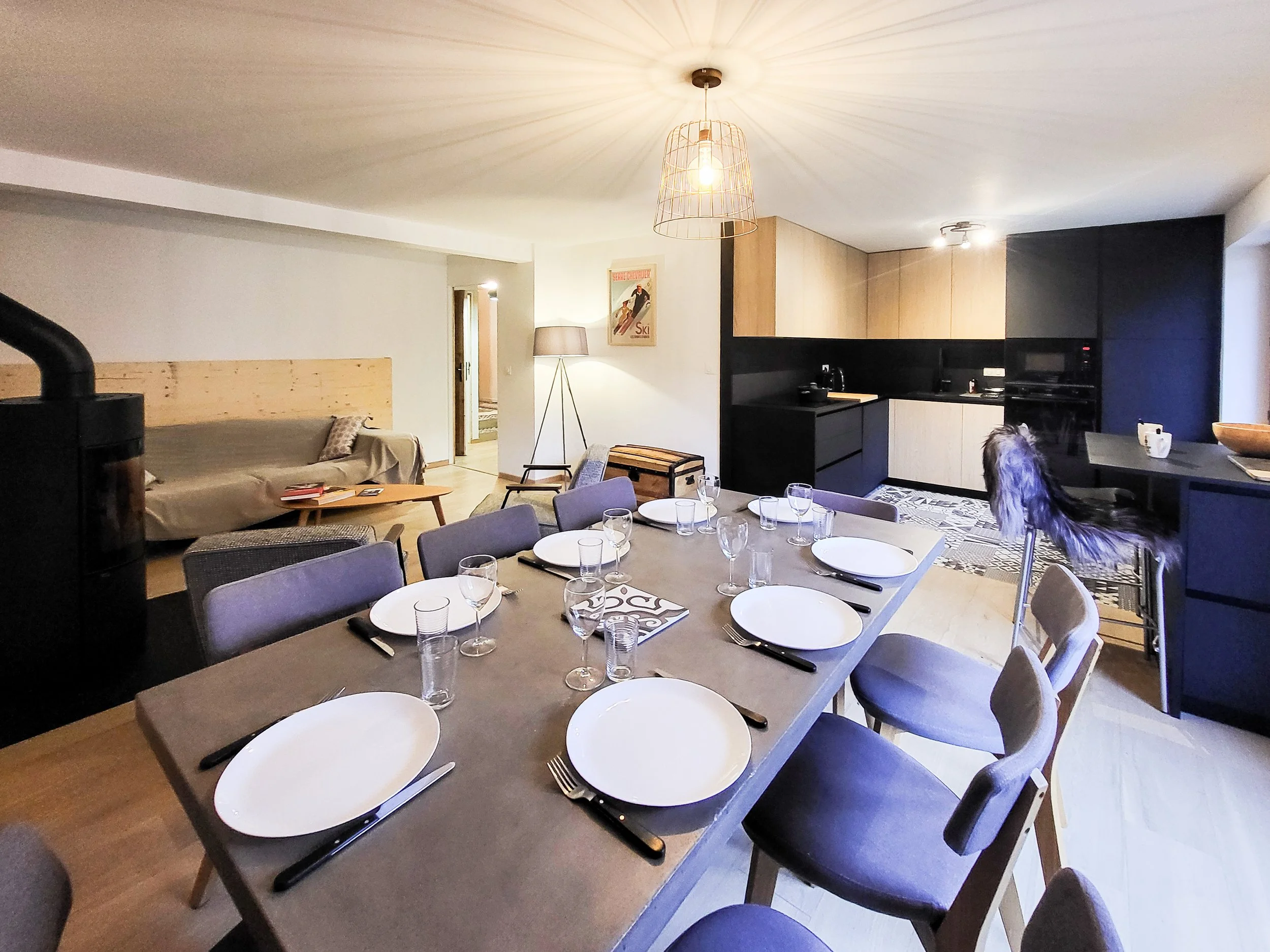 A modern dining room with a large rectangular table set with white plates, glasses, and cutlery, surrounded by upholstered chairs. The room features a kitchen with black and wooden cabinets, a sofa with a wooden headboard, a standing lamp, and a pain