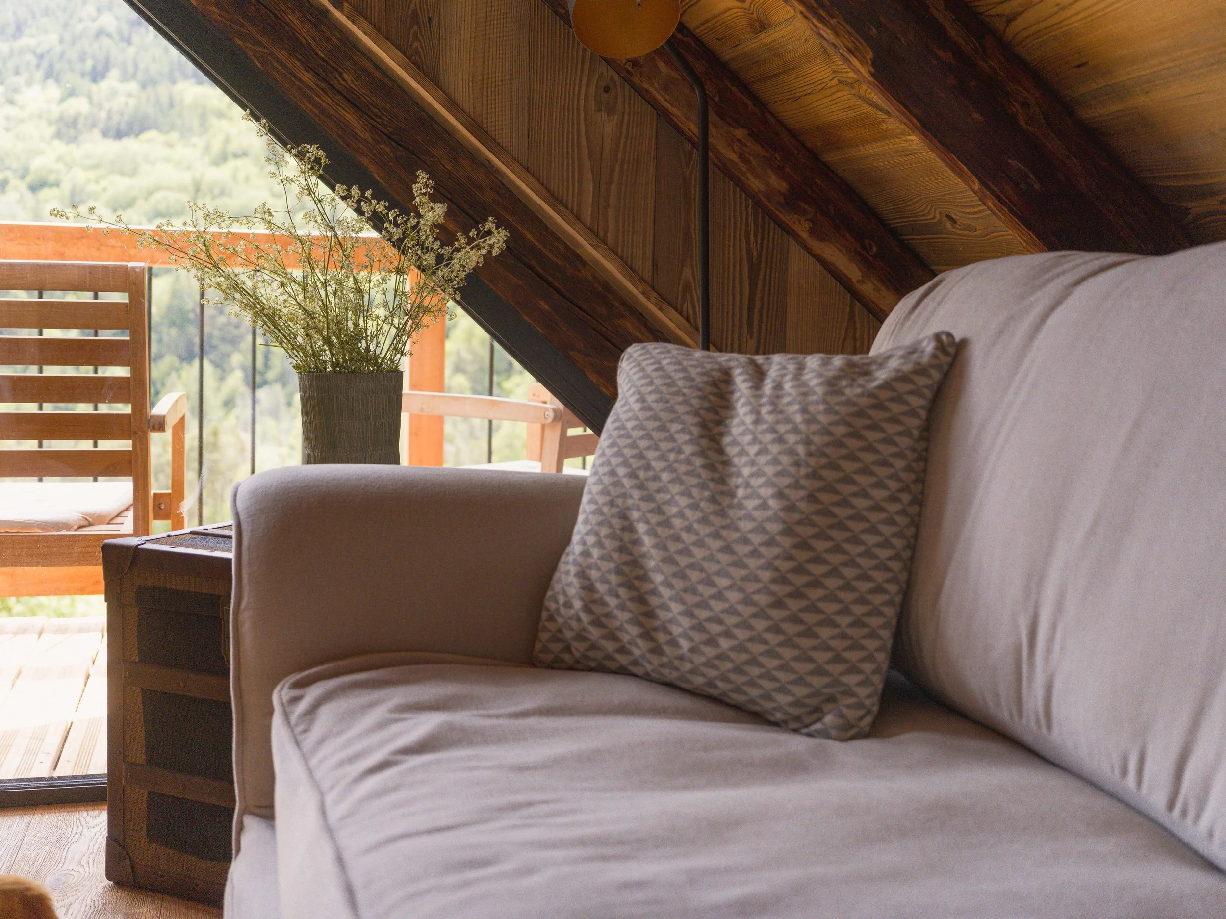 Une chambre mansardée chaleureuse en bois, avec un plafond en pente, où trône un lit orné d'oreillers à motifs, ainsi qu'un petit balcon donnant sur un paysage verdoyant avec les montagnes en arrière-plan, agrémenté d'un vase rempli de délicates fleurs blanches.