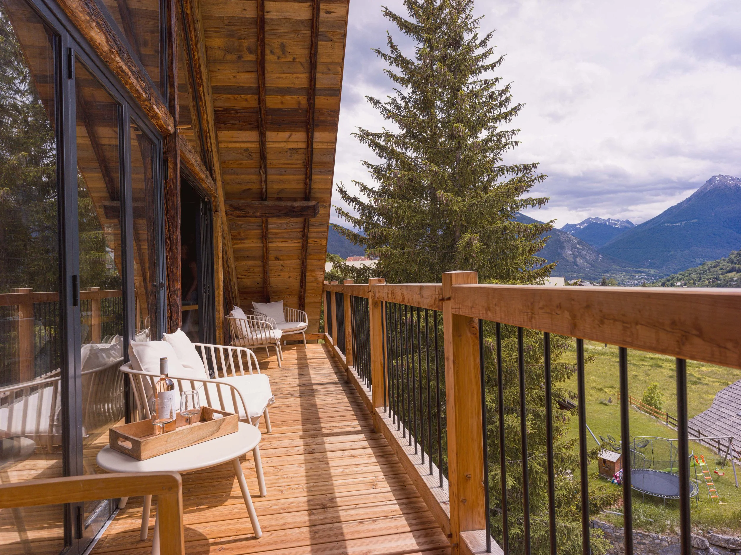 Un balcon en bois avec des chaises blanches et un plateau de boissons, offrant une vue panoramique sur les pins et les montagnes sous un ciel nuageux.