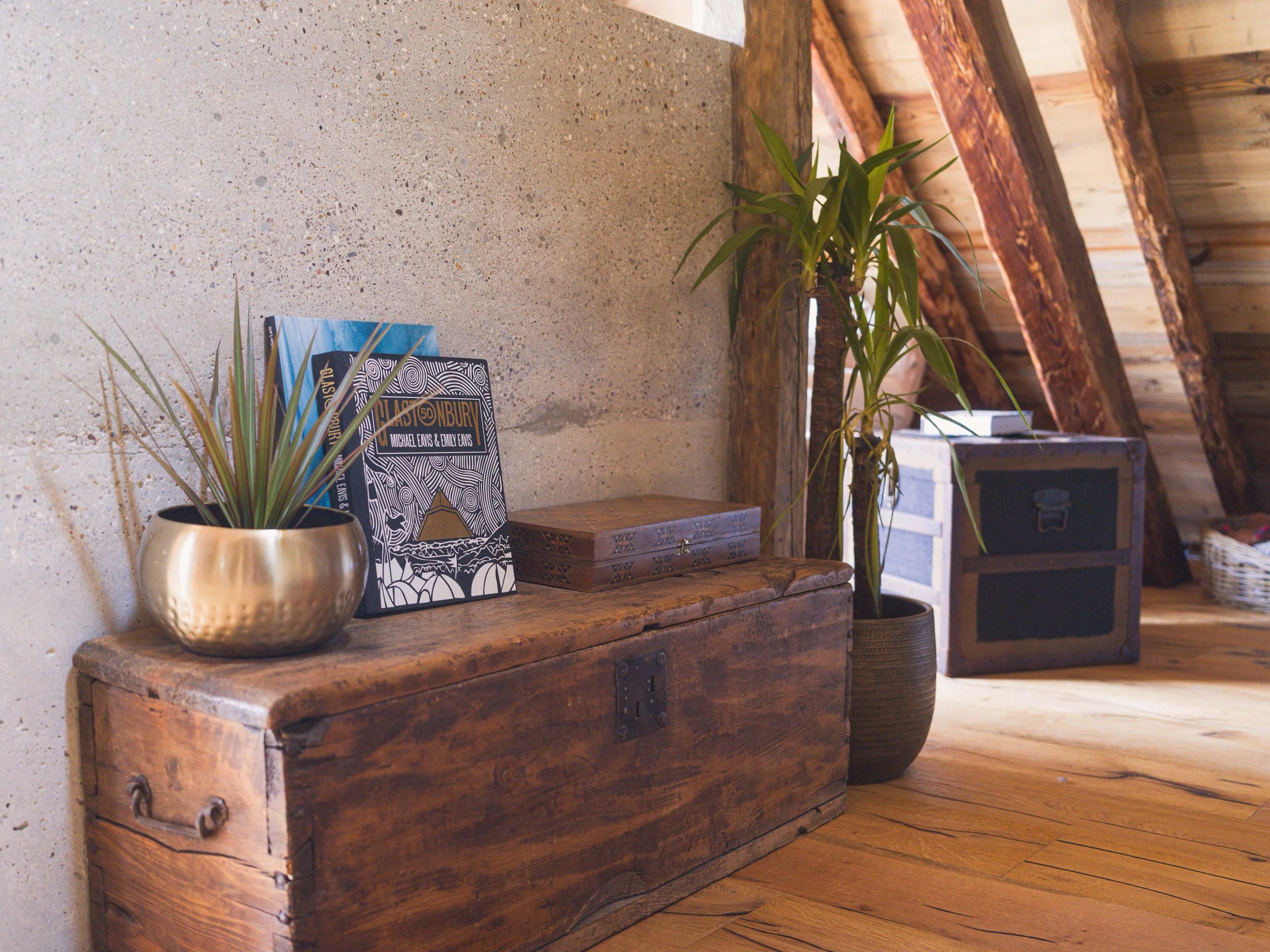 Un coffre en bois muni de poignées métalliques, une plante en pot, une brochure et une boîte décorative posée dessus, le tout dans un intérieur rustique avec des poutres apparentes et un parquet.
