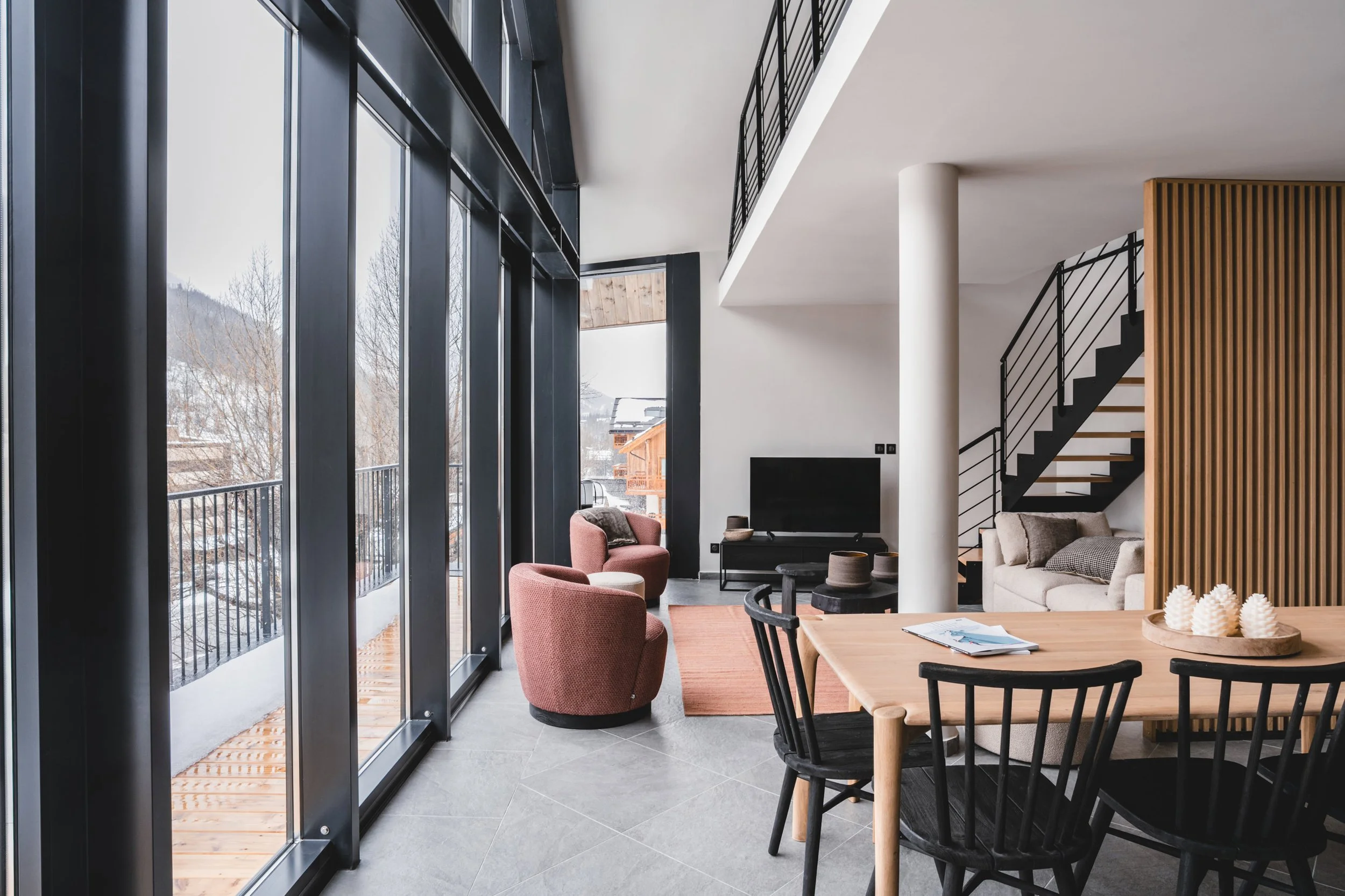 Interior of a modern living space with large glass windows, a dining table with black and wooden chairs, a sitting area with pink armchairs and a sofa, a flat-screen TV, and a staircase with black railing.