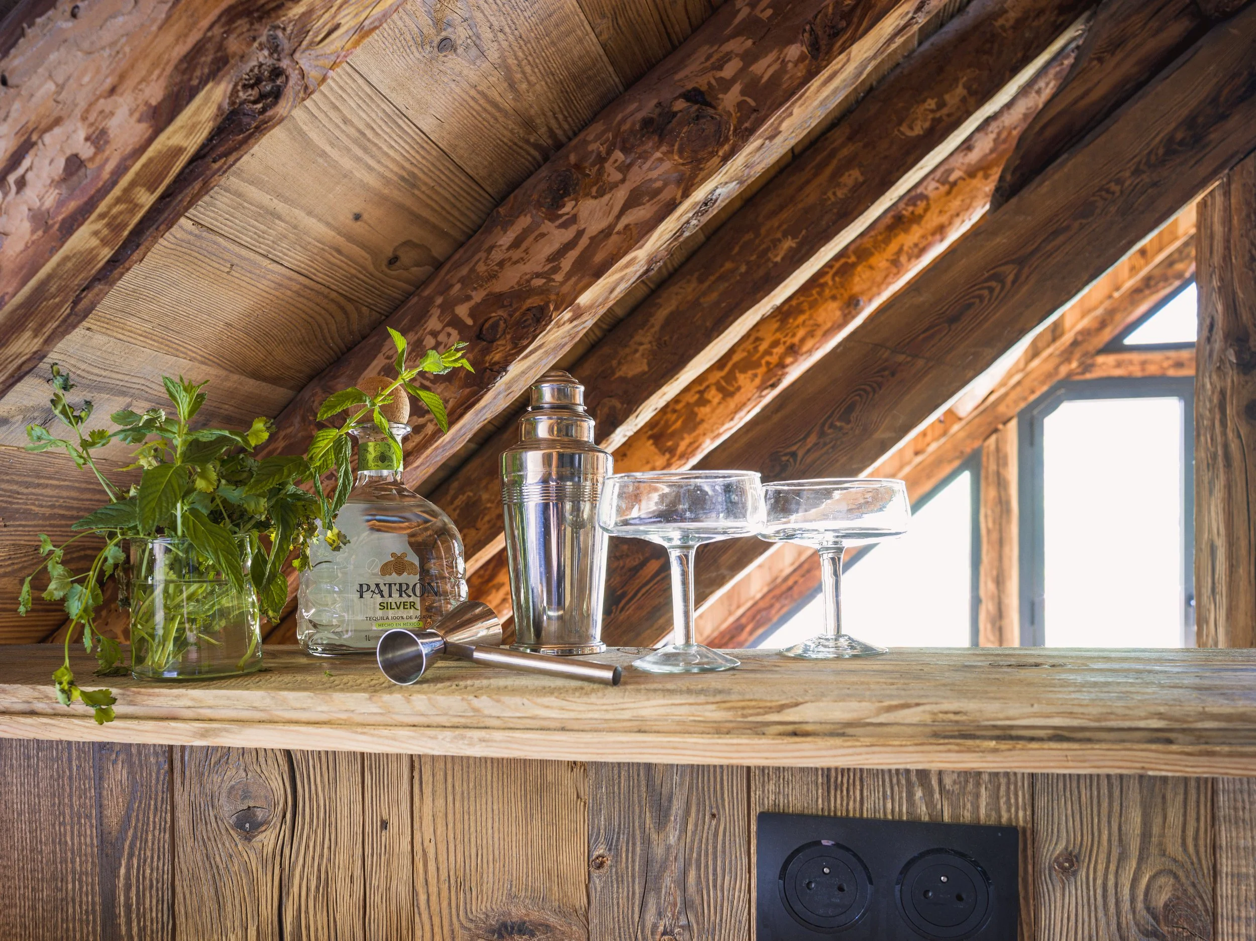Gros plan sur une étagère rustique en bois où sont posés un bocal en verre contenant des feuilles de menthe, une bouteille de tequila Patrón, quelques verres à shot et un shaker en métal, le tout devant un plafond en pente en bois percé d'une fenêtre.