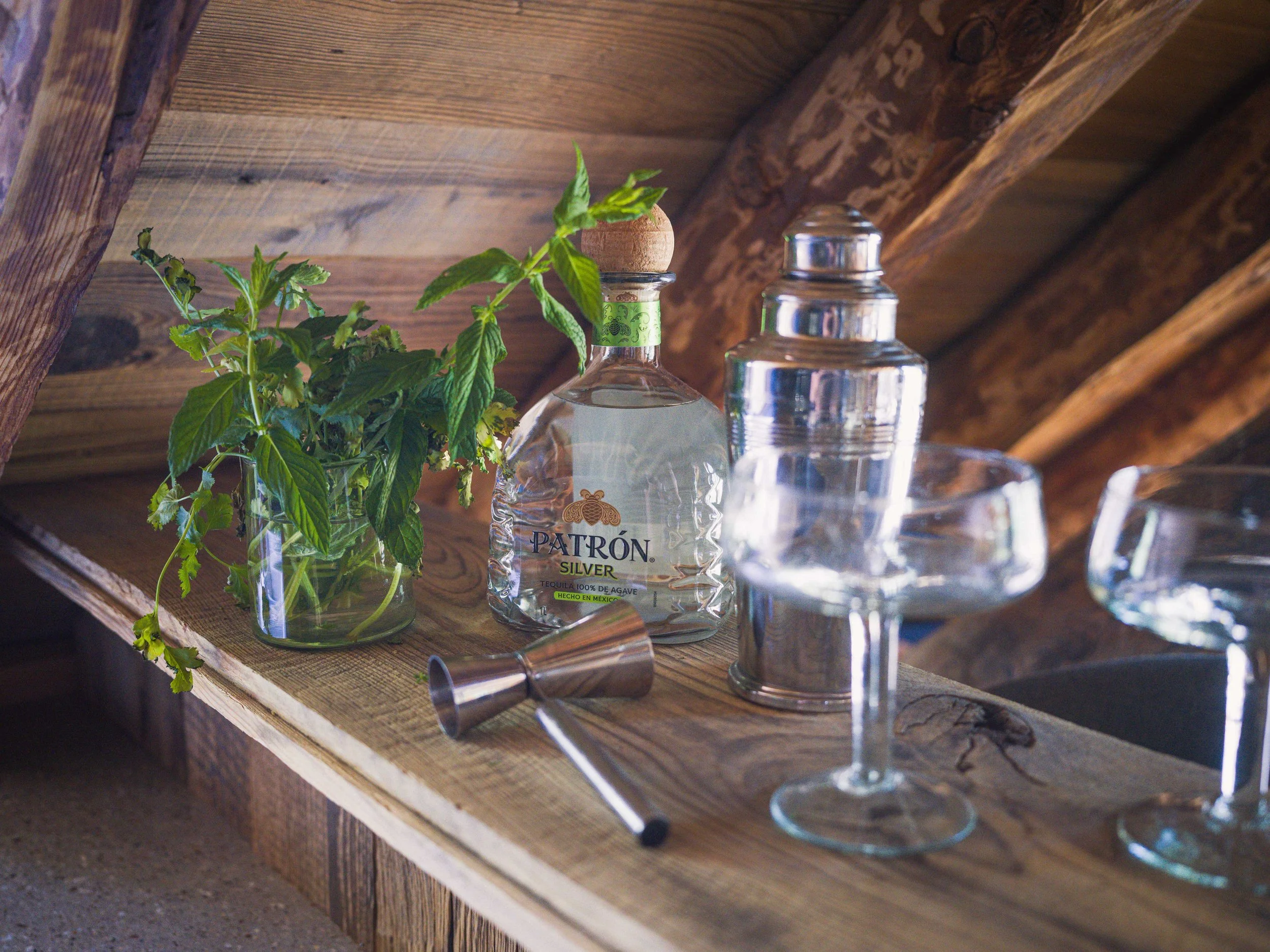 Une étagère en bois sur laquelle se trouvent une bouteille de tequila Patrón Silver, un petit verre, un doseur en argent et un bouquet de feuilles de menthe fraîche dans un récipient en verre.