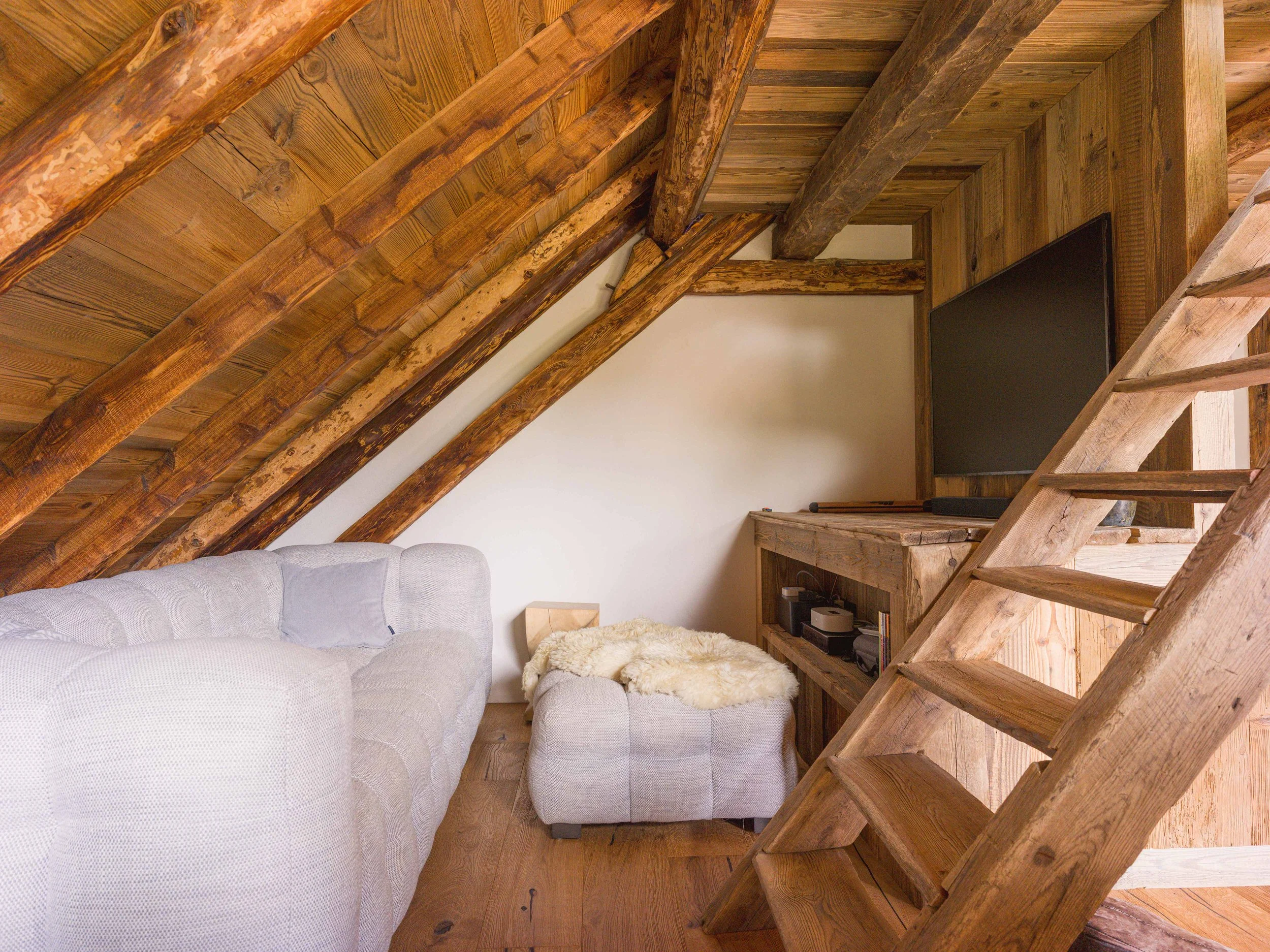 Vue d'un grenier confortable avec des poutres et un plafond apparents, un canapé de couleur claire recouvert d'un plaid moelleux, ainsi qu'une bibliothèque décorative sur laquelle trône un téléviseur à écran plat, le tout sur un mur en bois.