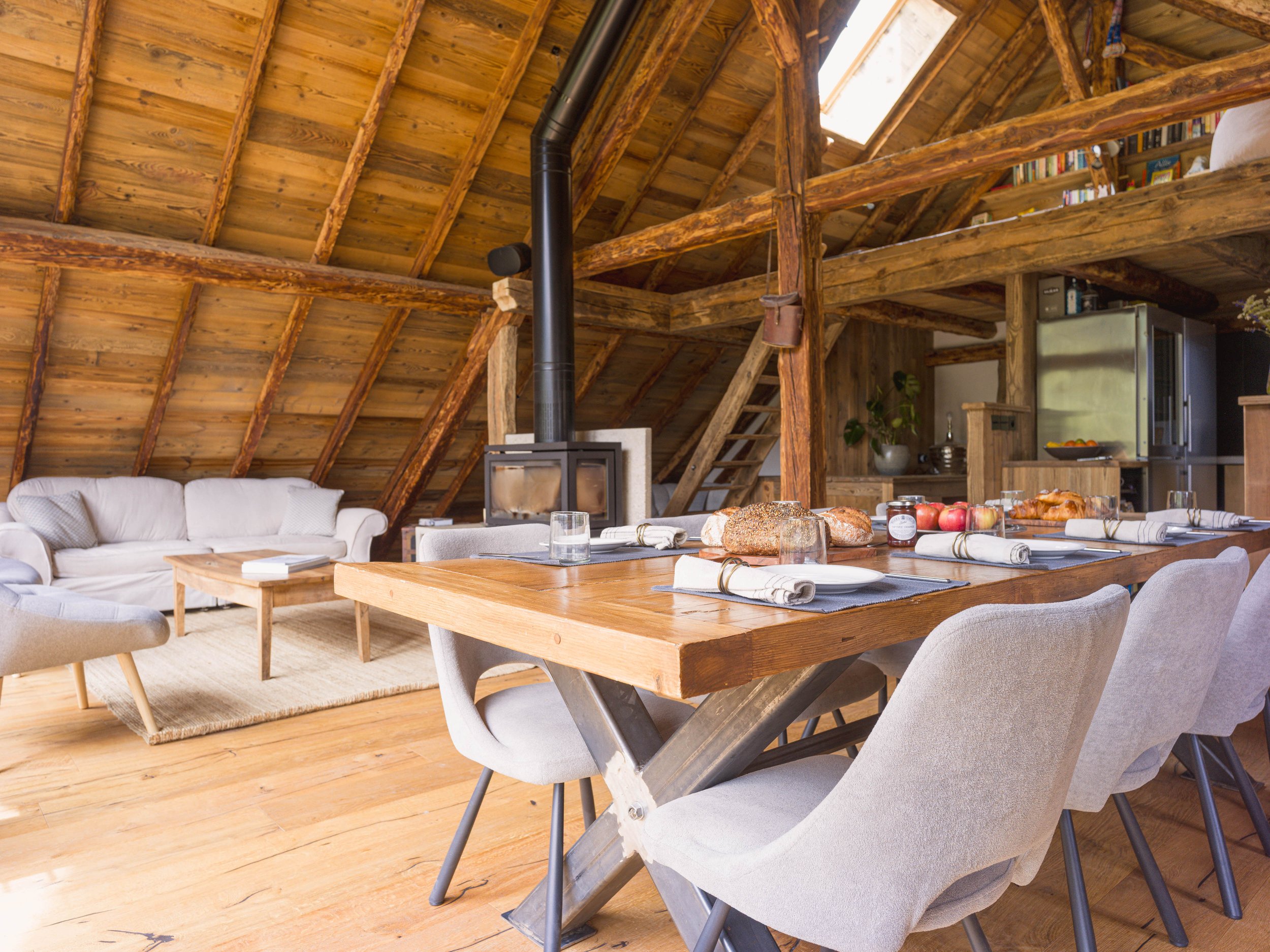 Intérieur d'un grenier en bois chaleureux, avec une table à manger, des chaises, un canapé, une cheminée en pierre et des étagères, baigné par la lumière naturelle provenant d'une lucarne.