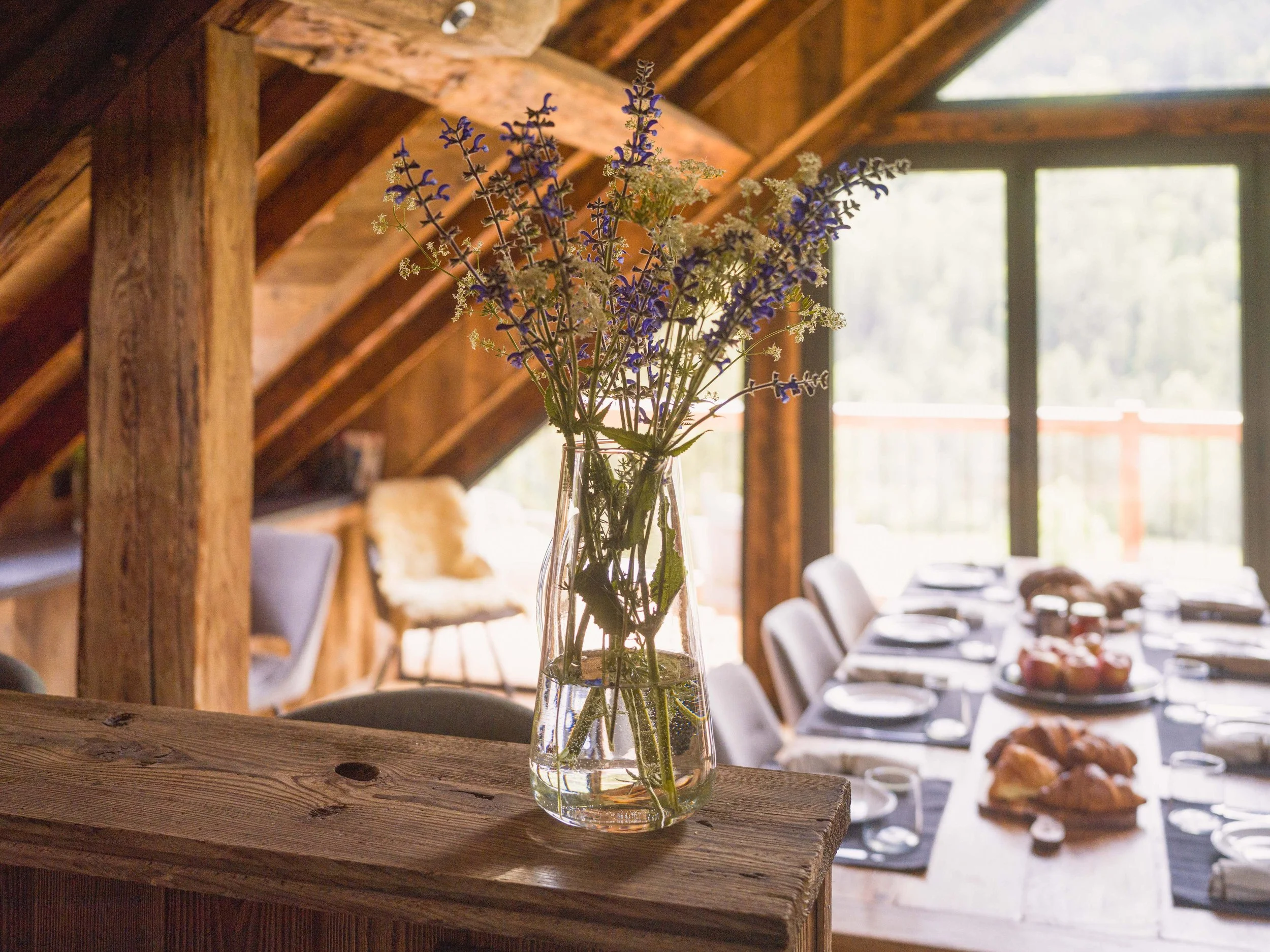 Un vase en verre transparent contenant des fleurs violettes et blanches, posé sur un rebord en bois dans un intérieur rustique, avec la lumière naturelle qui pénètre par les fenêtres en arrière-plan.