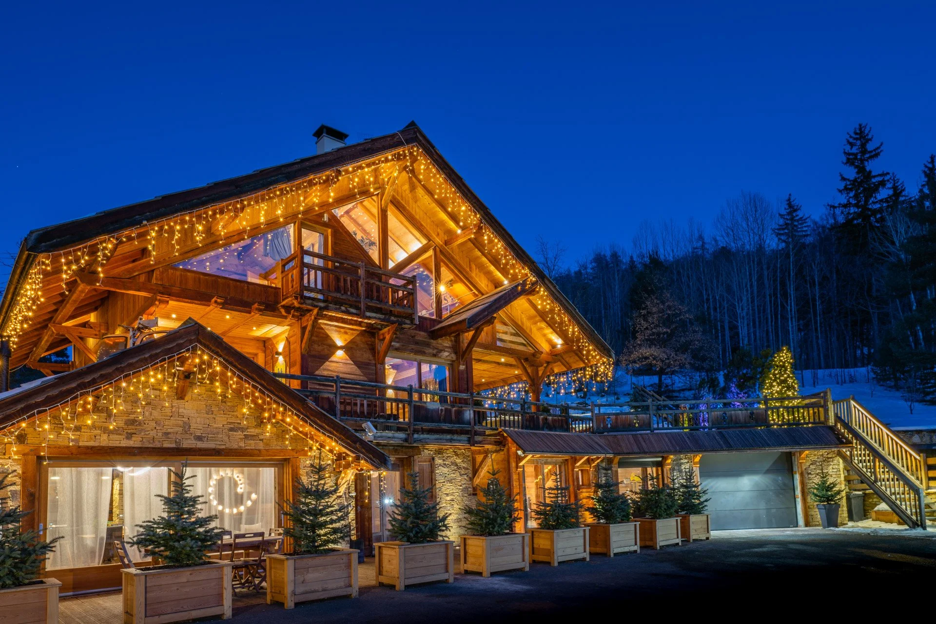 A multi-story wooden house decorated with Christmas lights at dusk, surrounded by pine trees and snow, with a staircase on the right side.