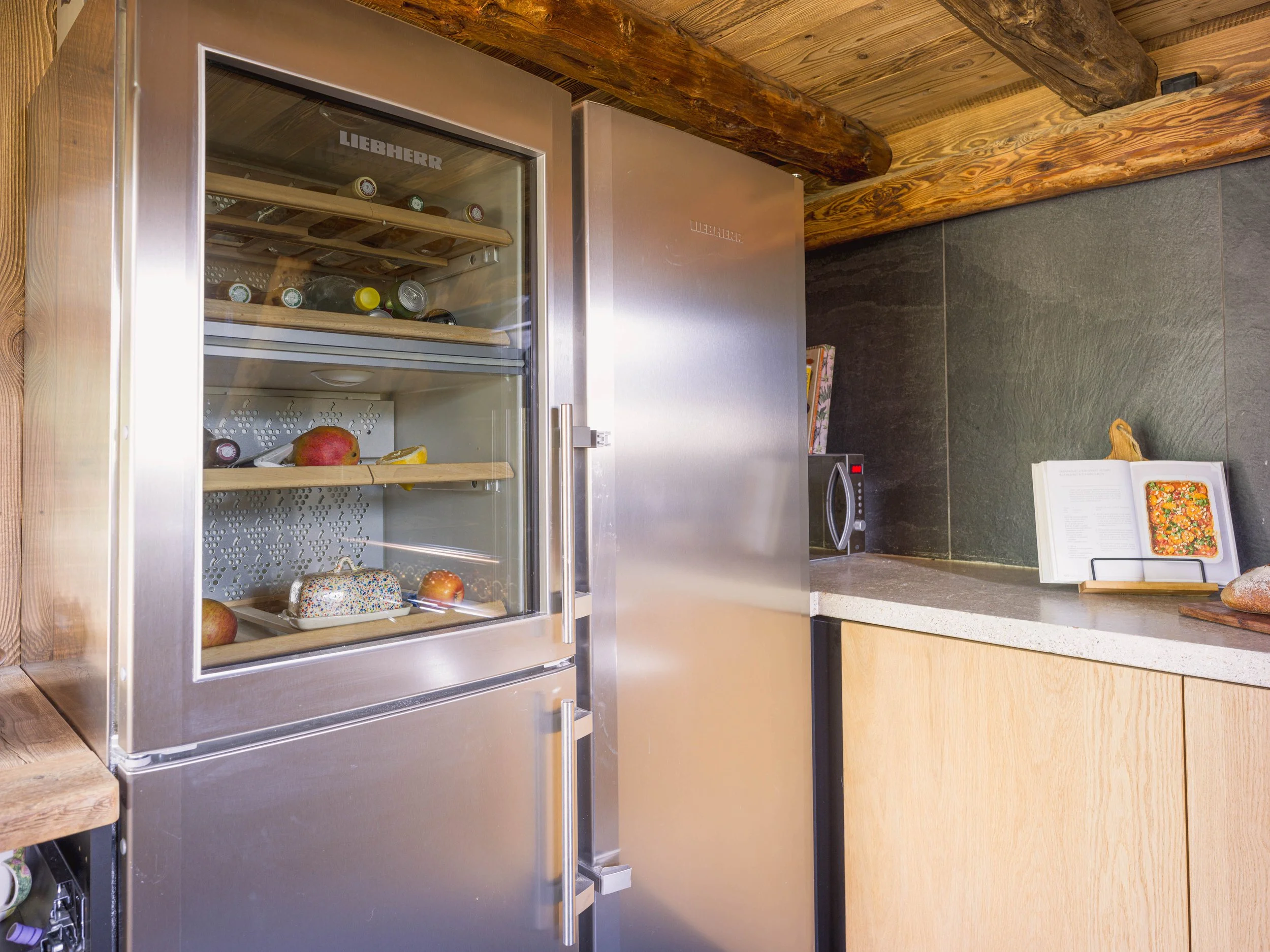 Une cave à vin Liebherr en acier inoxydable dotée d'une porte vitrée, remplie de bouteilles de vin et d'autres boissons, posée sur un plan de travail en bois dans une cuisine rustique aux poutres apparentes et aux murs carrelés de couleur sombre.