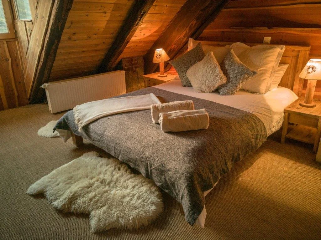 A cozy bedroom with a sloped wooden ceiling, a large bed with multiple pillows, a blanket, and two bedside tables with lamps, a fluffy rug, and a cream-colored sheepskin on the floor.