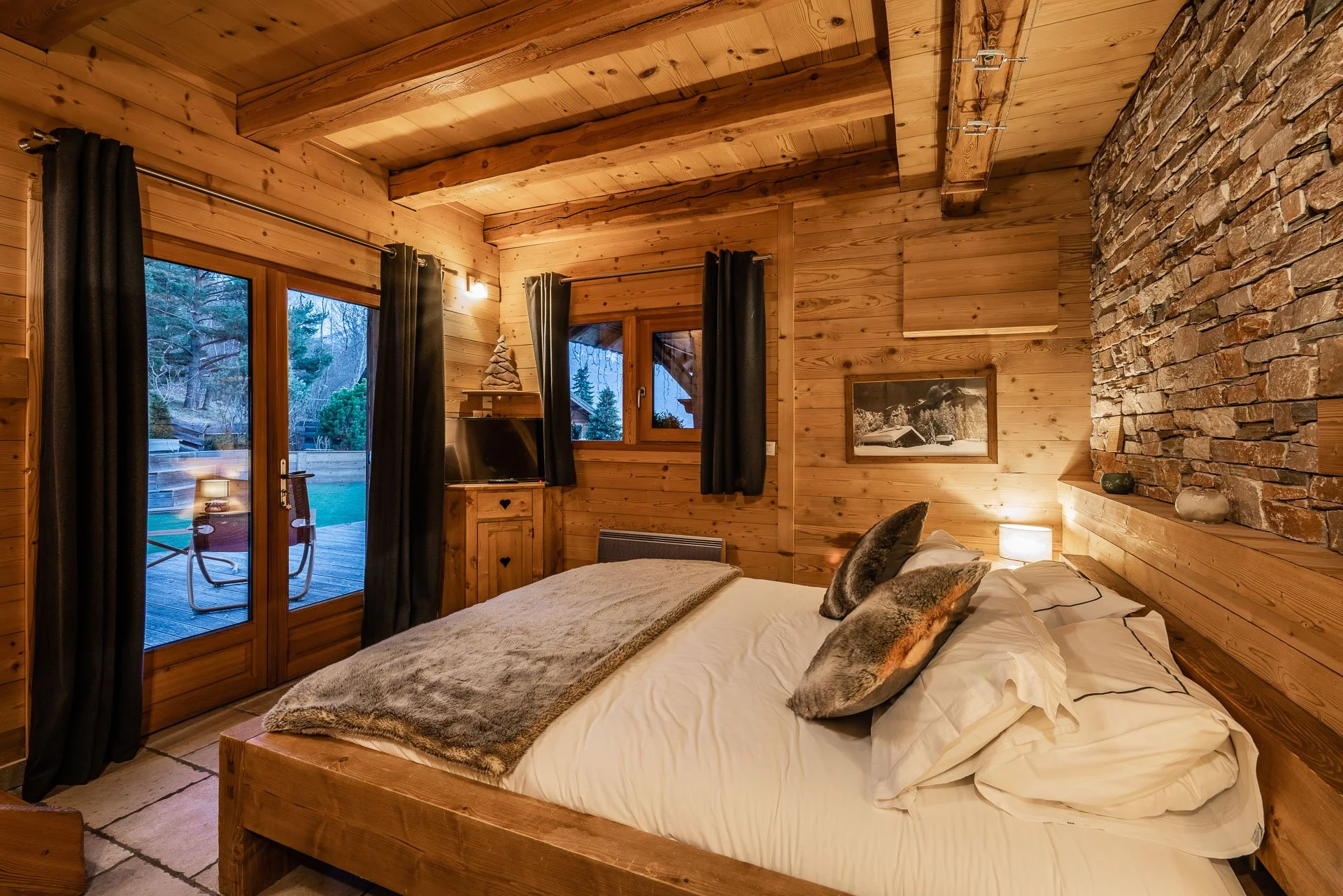 Cozy bedroom with wooden walls, a large bed with white linens and two furry pillows, a small nightstand with a lamp, black curtains, and a sliding glass door leading to an outdoor deck. There is a window and a framed black-and-white picture on the wa