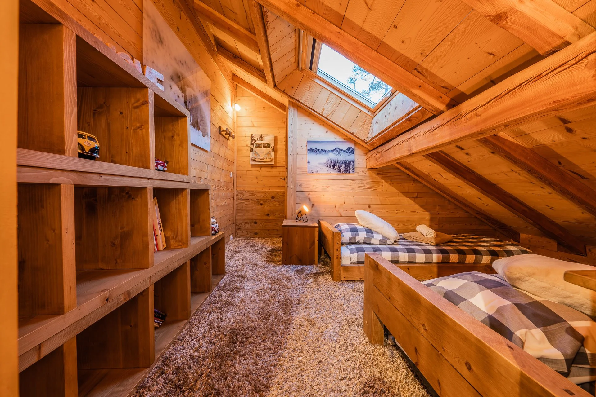 A cozy wooden attic bedroom with two beds, a skylight window, a bookshelf, and mountain landscape artwork.