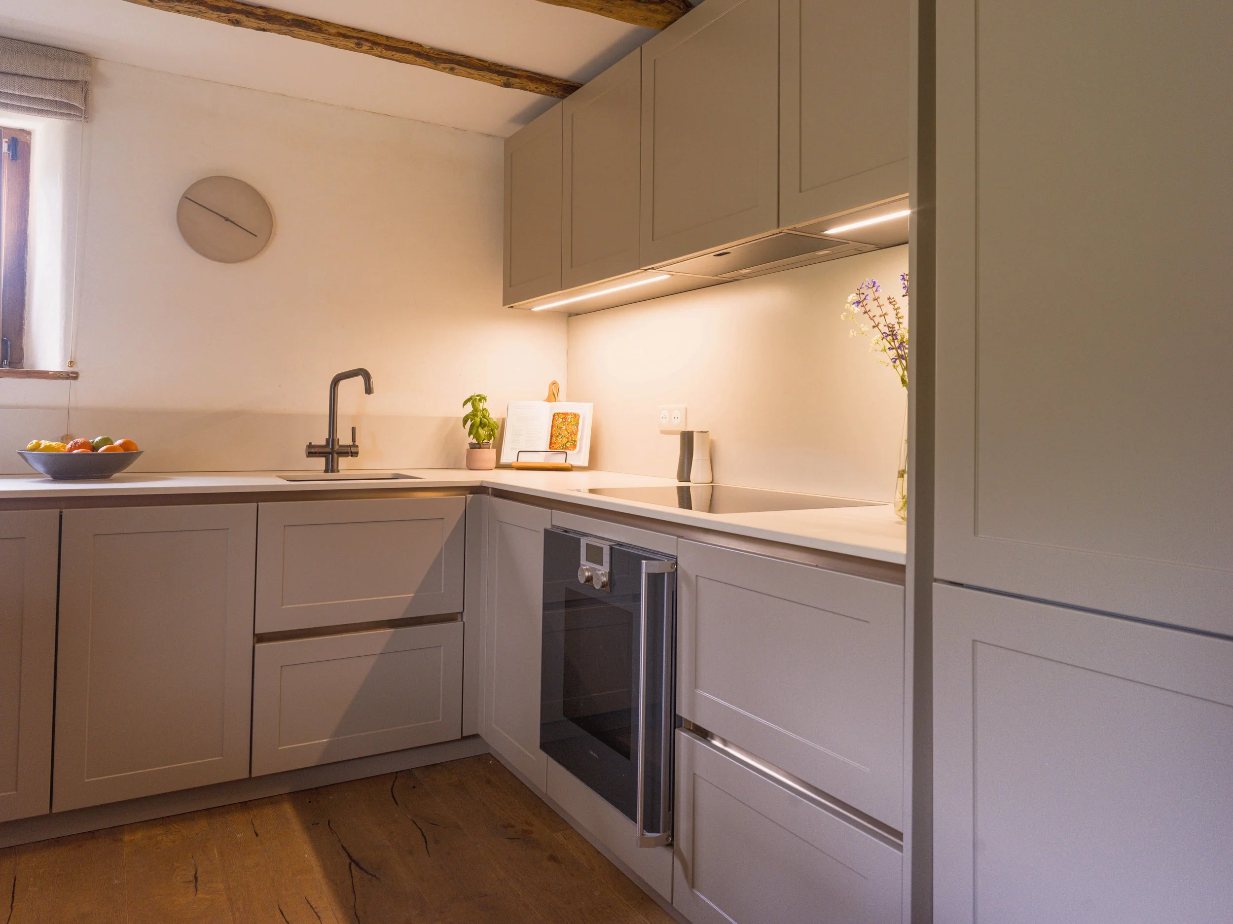 Modern kitchen with gray cabinetry, white countertops, a black faucet, a fruit bowl, a plant, a book, and a built-in oven under upper cabinets. There is a round wall clock and a window with a Roman shade.