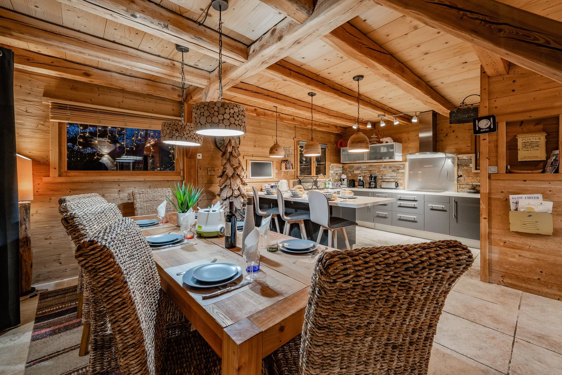 Wood-paneled kitchen and dining area with a large wooden table, woven chairs, modern gray cabinetry, a kitchen island, hanging pendant lights, and a window with night sky view.