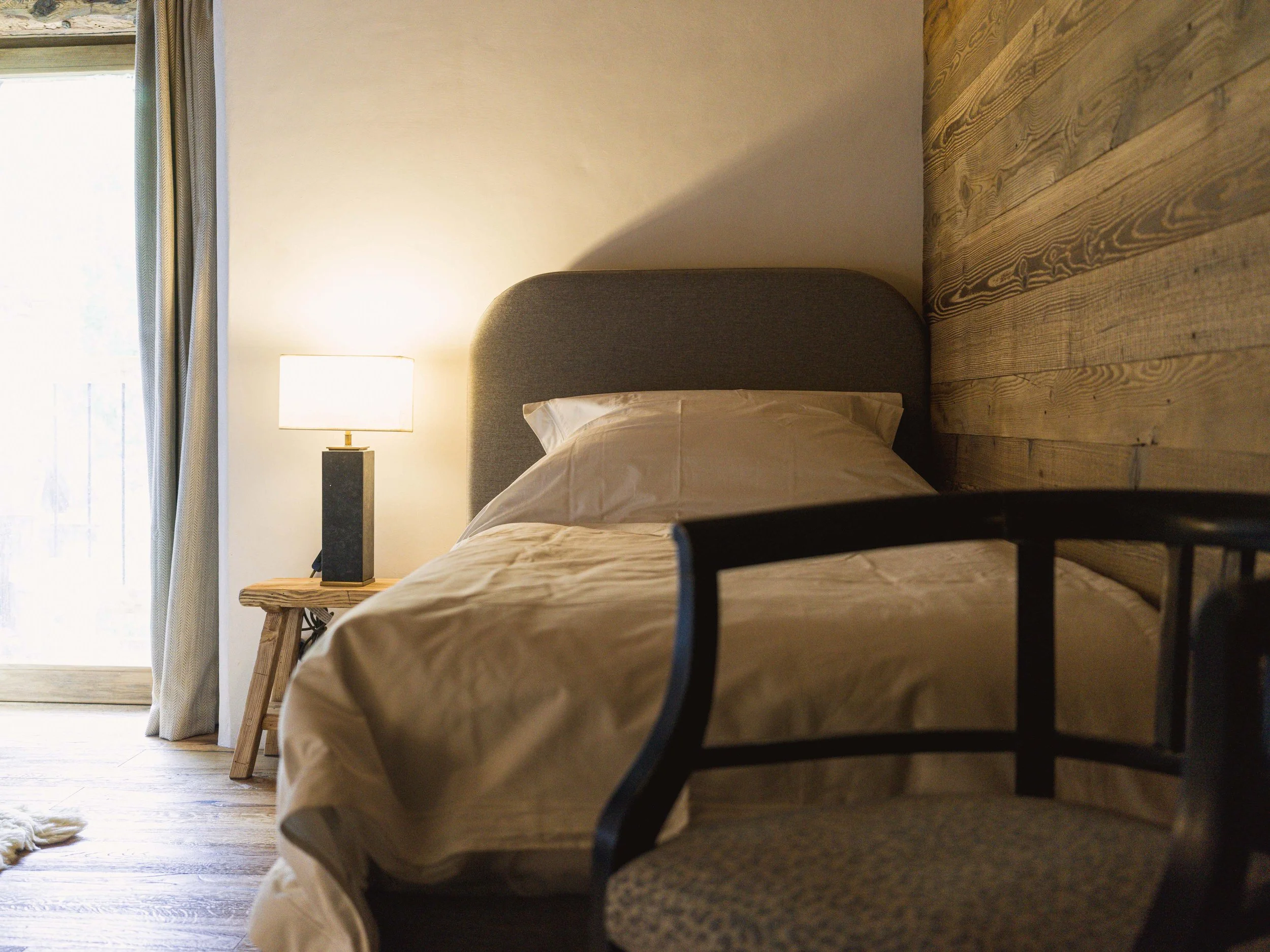 Une chambre avec un lit, une table de chevet sur laquelle se trouve une lampe, un parquet et une fenêtre avec des rideaux.