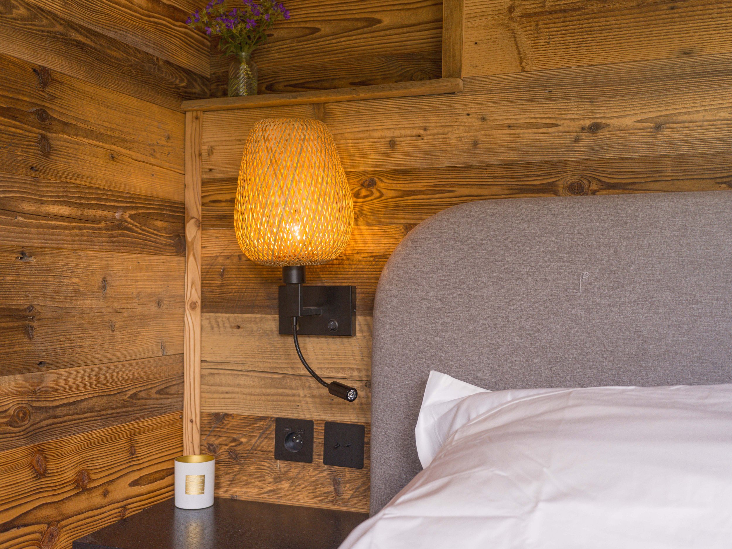 Gros plan sur une chambre aux murs en bois, avec un lit gris recouvert de draps blancs, une table de chevet noire sur laquelle se trouve une bougie, une lampe fixée au mur et une petite étagère sur laquelle est posé un vase contenant des fleurs violettes.