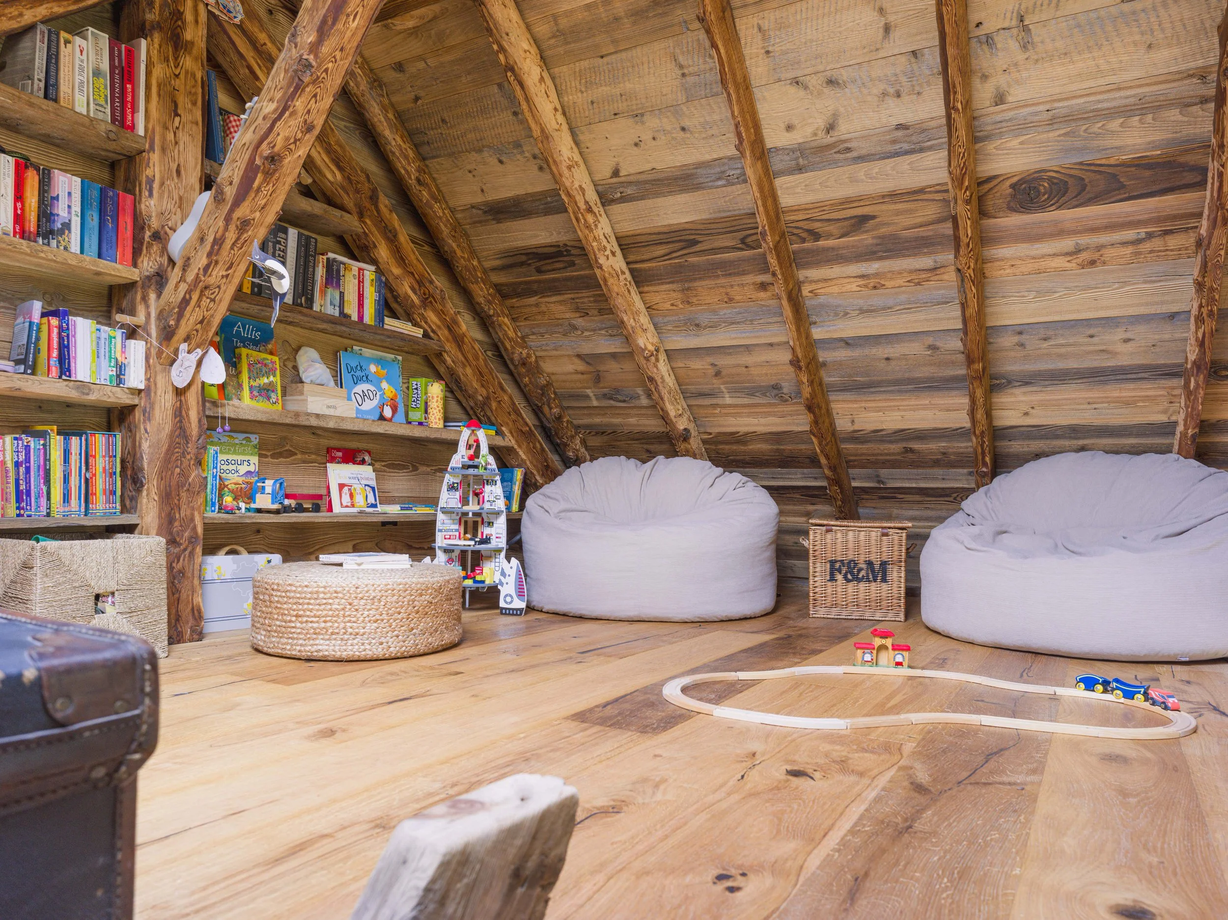 Une chambre mansardée chaleureuse avec des murs et un plafond en bois, une bibliothèque remplie de livres, des poufs moelleux, une petite table en bois et un circuit de train avec des petits trains sur le sol.