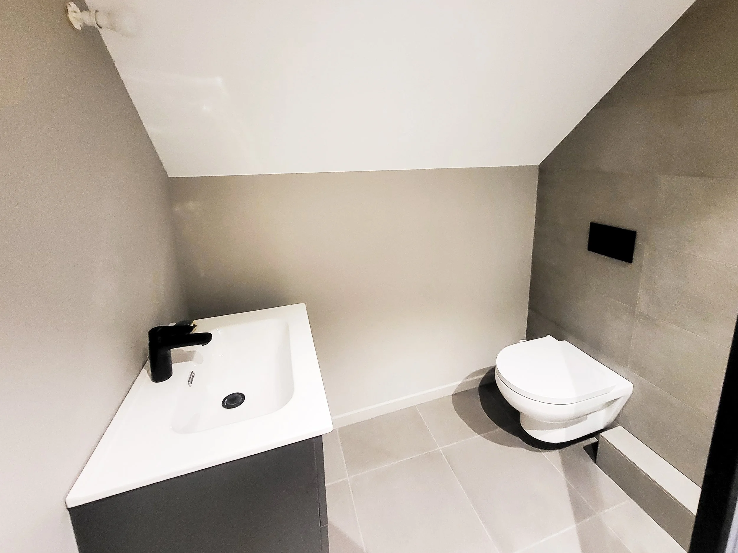 Modern bathroom with a white wall-mounted toilet and a black faucet sink on a gray cabinet, beige walls, large gray tiles on the floor, and a black flush panel.