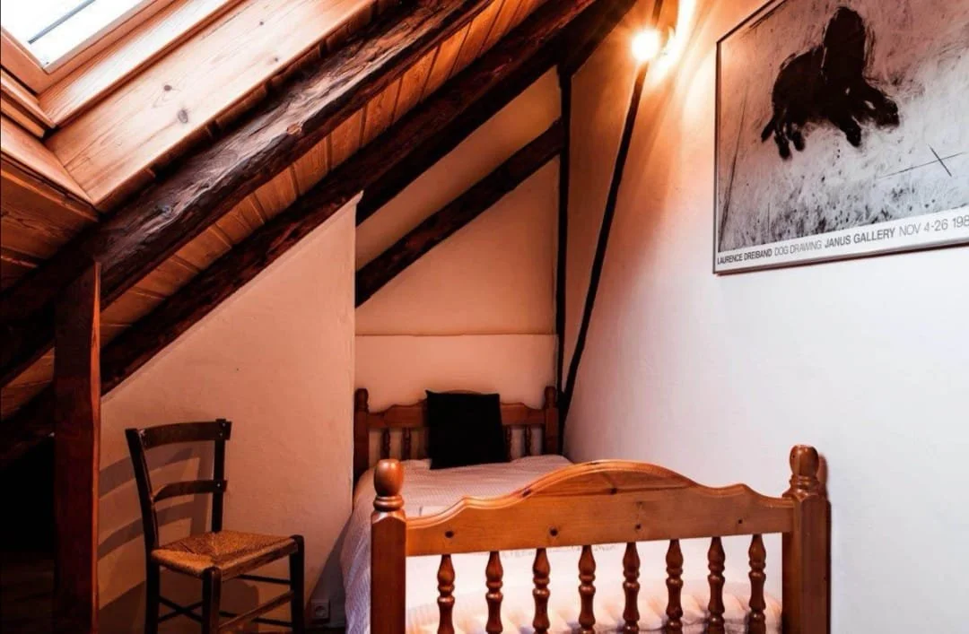 A cozy attic bedroom with a wooden bed, a black pillow, an upright chair, and a framed dog drawing on the wall beneath a sloped wooden ceiling with skylight.