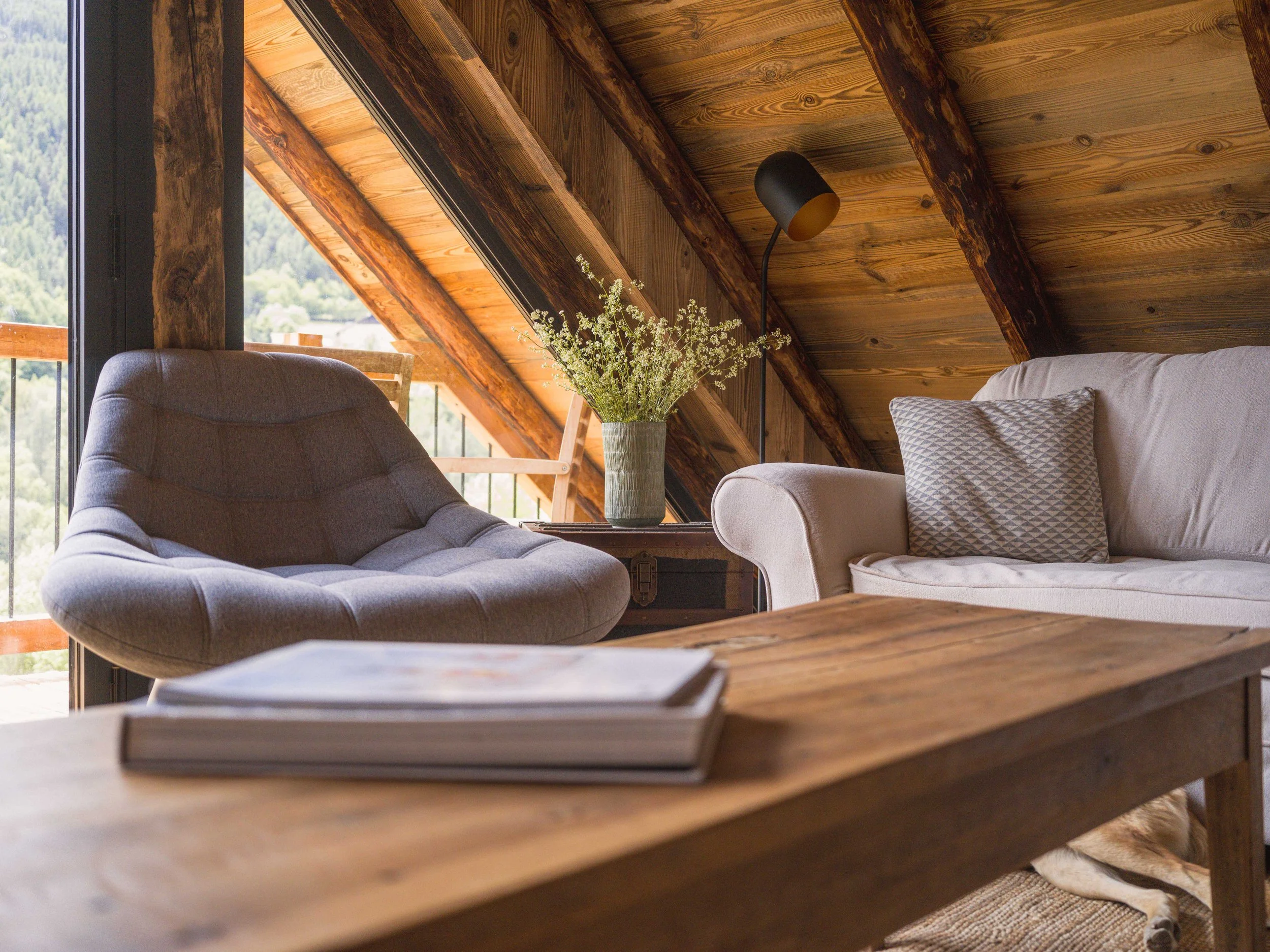 Salon avec un plafond en bois en pente, une grande fenêtre donnant sur la montagne, un fauteuil gris, un canapé beige orné d'un coussin à motifs, une applique murale noire et une table basse en bois sur laquelle sont posés des livres et un vase de fleurs blanches.
