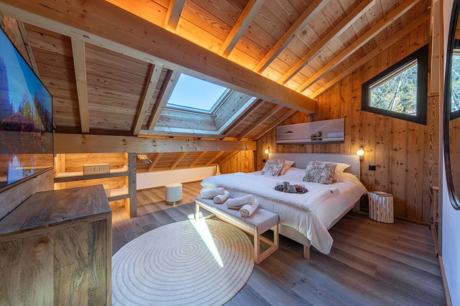 A cozy bedroom with wooden walls and ceiling, skylight, and windows showing trees outside, featuring a white bed with pillows and a tray of breakfast items, a round rug, and modern furniture.