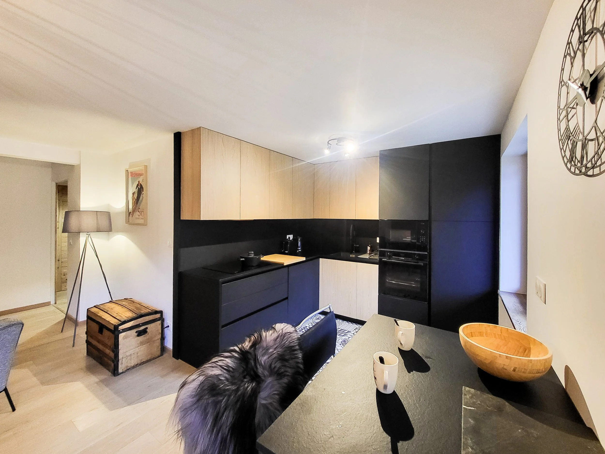 Modern kitchen with black and wooden cabinets, a black countertop, and a window. There is a counter with two mugs and a wooden bowl. A dog with gray fur is partially visible in front of the counter. A tall floor lamp and a framed picture are on the w