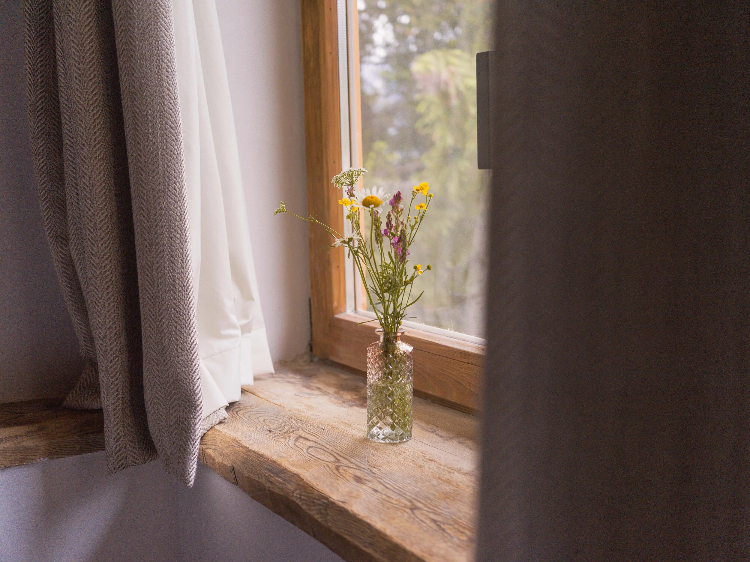 Un vase en verre transparent orné d'un motif en losanges, contenant un petit bouquet de fleurs des champs, posé sur un rebord de fenêtre en bois à côté des rideaux, avec une vue sur l'extérieur visible à travers la fenêtre.