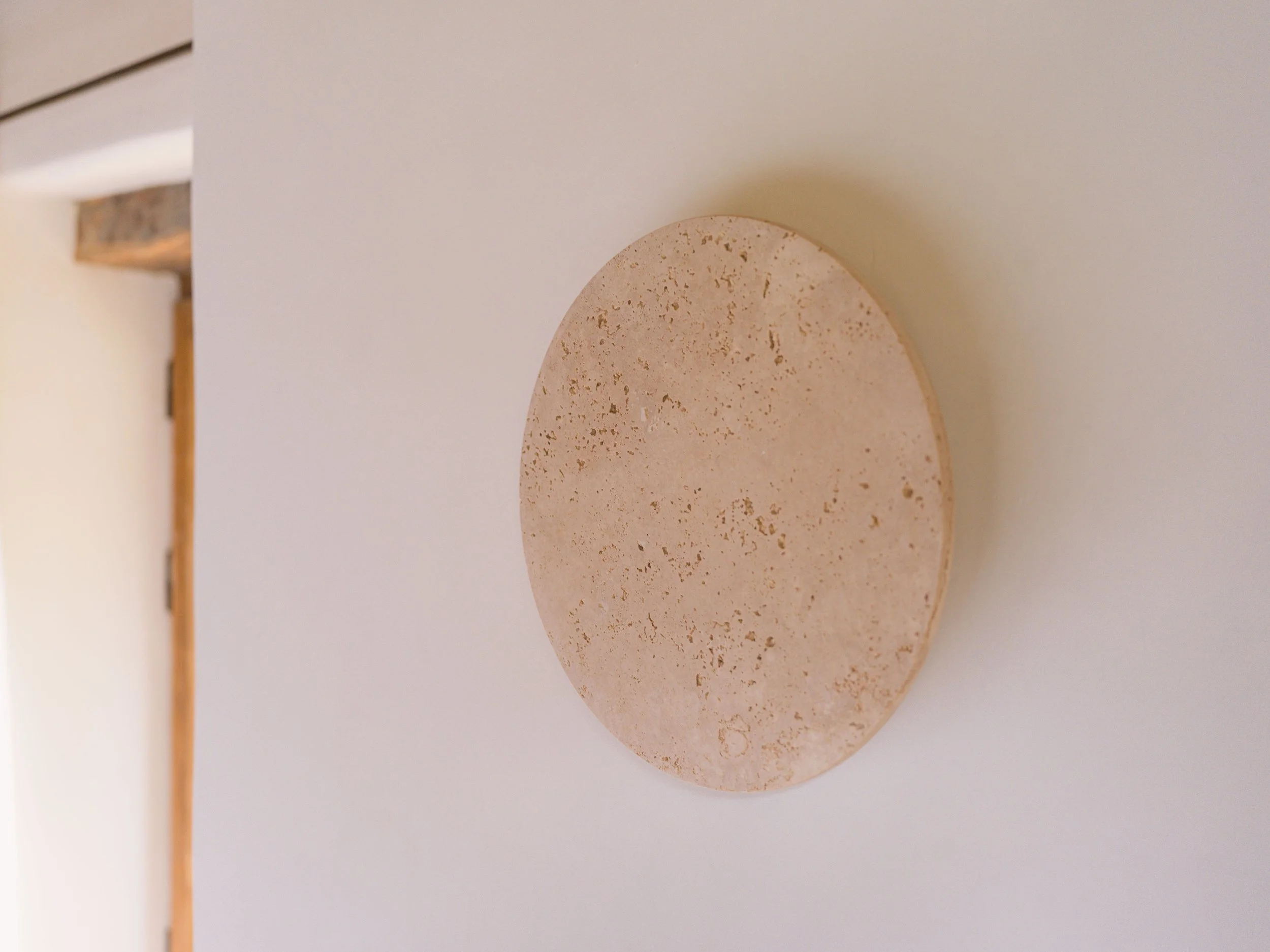 Gros plan sur un objet circulaire beige moucheté, peut-être un disque en pierre ou en céramique, posé sur une surface blanche avec un petit espace en haut, sur fond comportant des éléments beiges et marron.