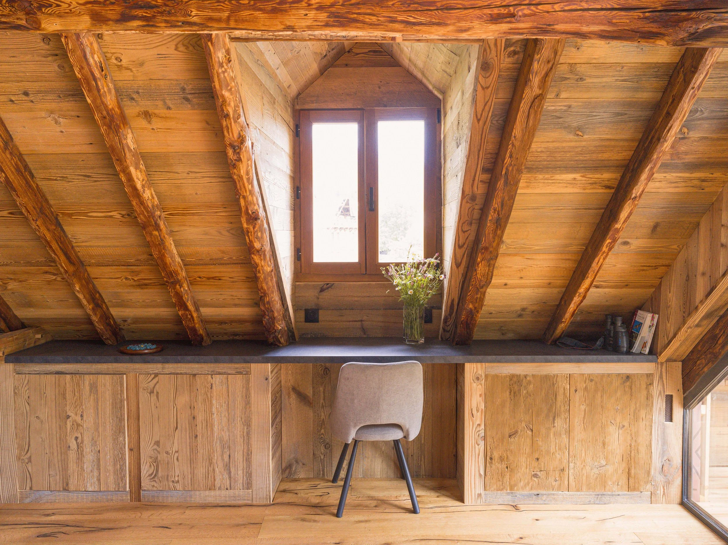 Vue intérieure d'un grenier confortable avec un plafond en bois, des fenêtres, un bureau, une chaise, un vase à fleurs et des étagères.