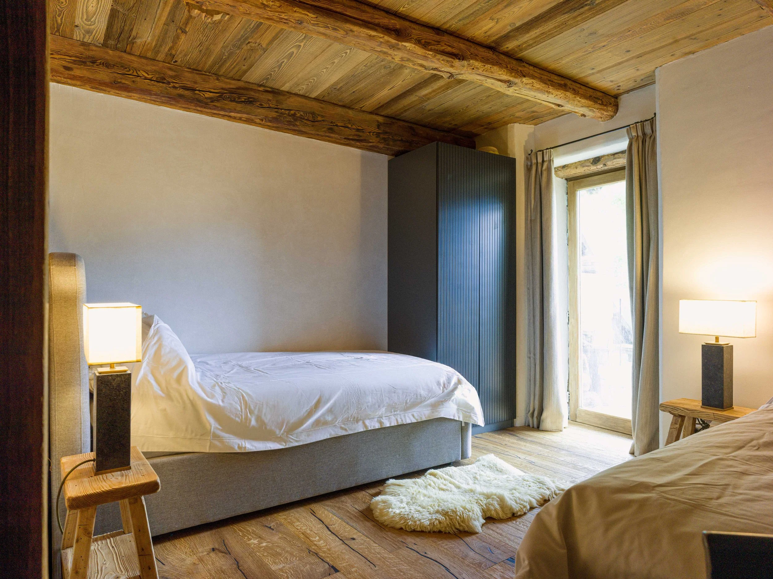 Une chambre avec un plafond et un sol en bois, comprenant un lit avec une parure de lit blanche, une petite table de chevet en bois avec une lampe, et une armoire noire près d'une grande fenêtre ornée de rideaux. Un tapis moelleux recouvre le sol.