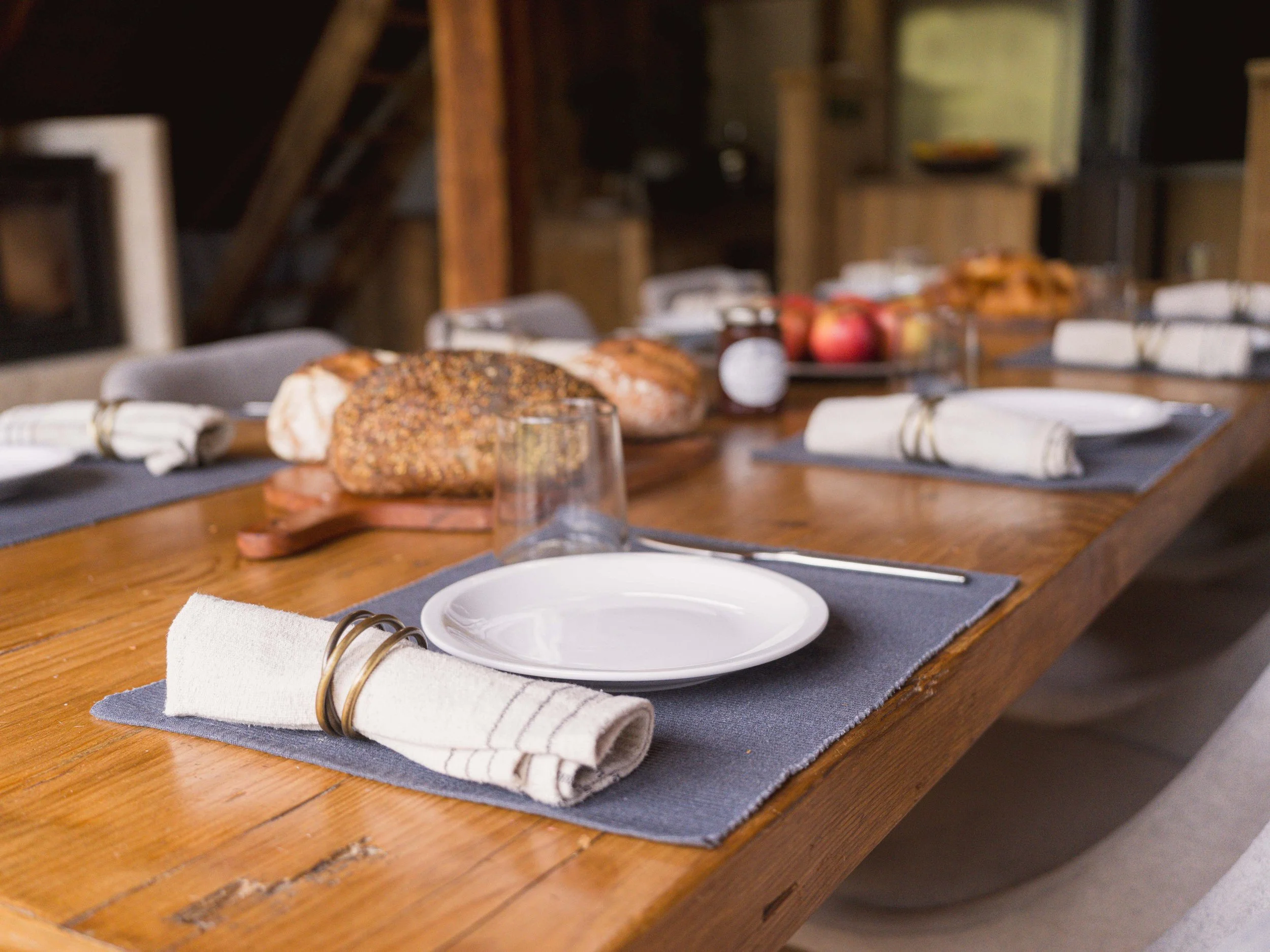 Une table à manger rustique en bois dressée pour un repas, avec des serviettes, des assiettes, des verres, du pain, des pommes et un pot de confiture, dans un intérieur chaleureux en bois.