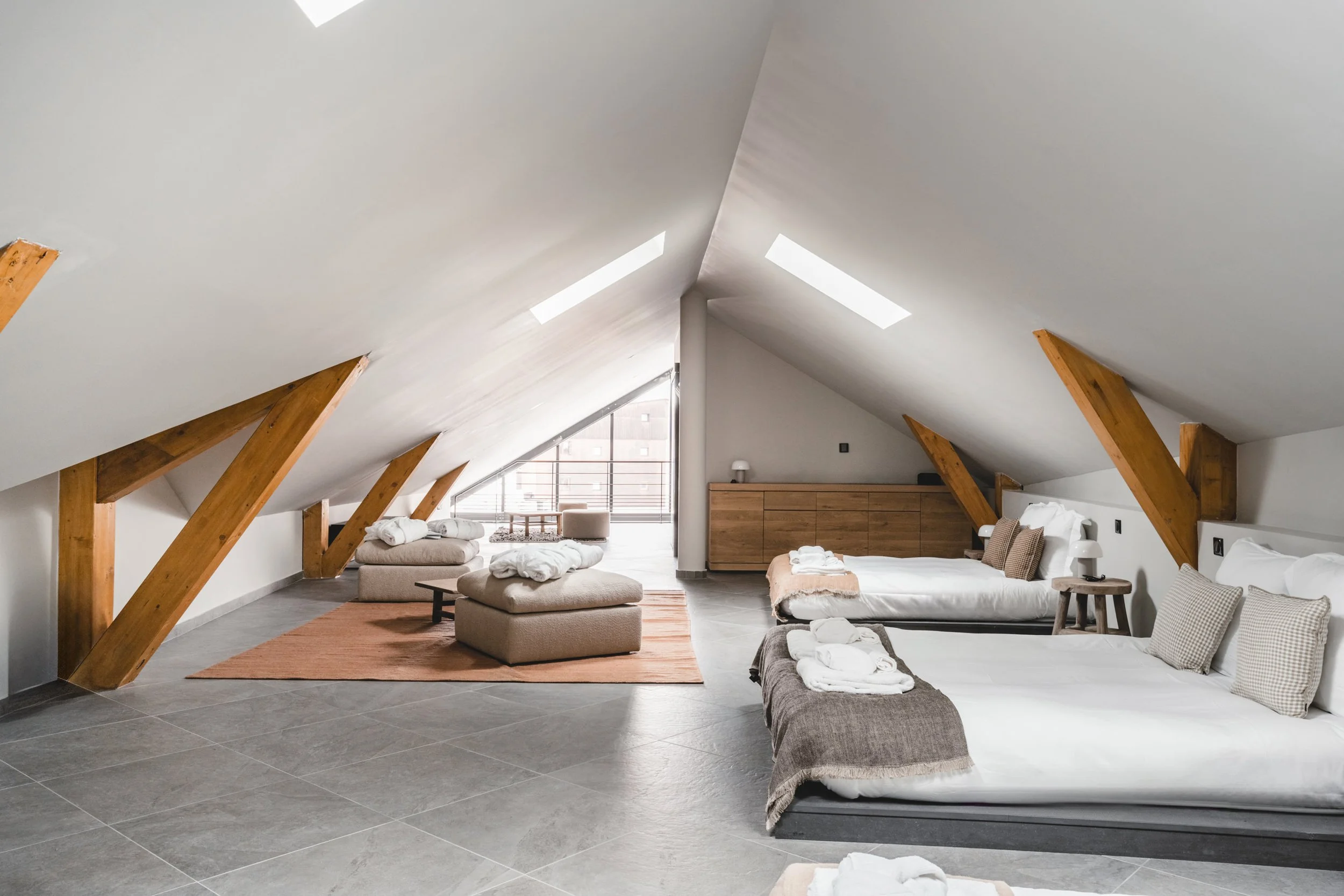 Spacious attic bedroom with sloped white ceiling, skylights, two beds with gray and beige bedding, small side tables, and a wooden dresser, illuminated by natural light from a balcony.