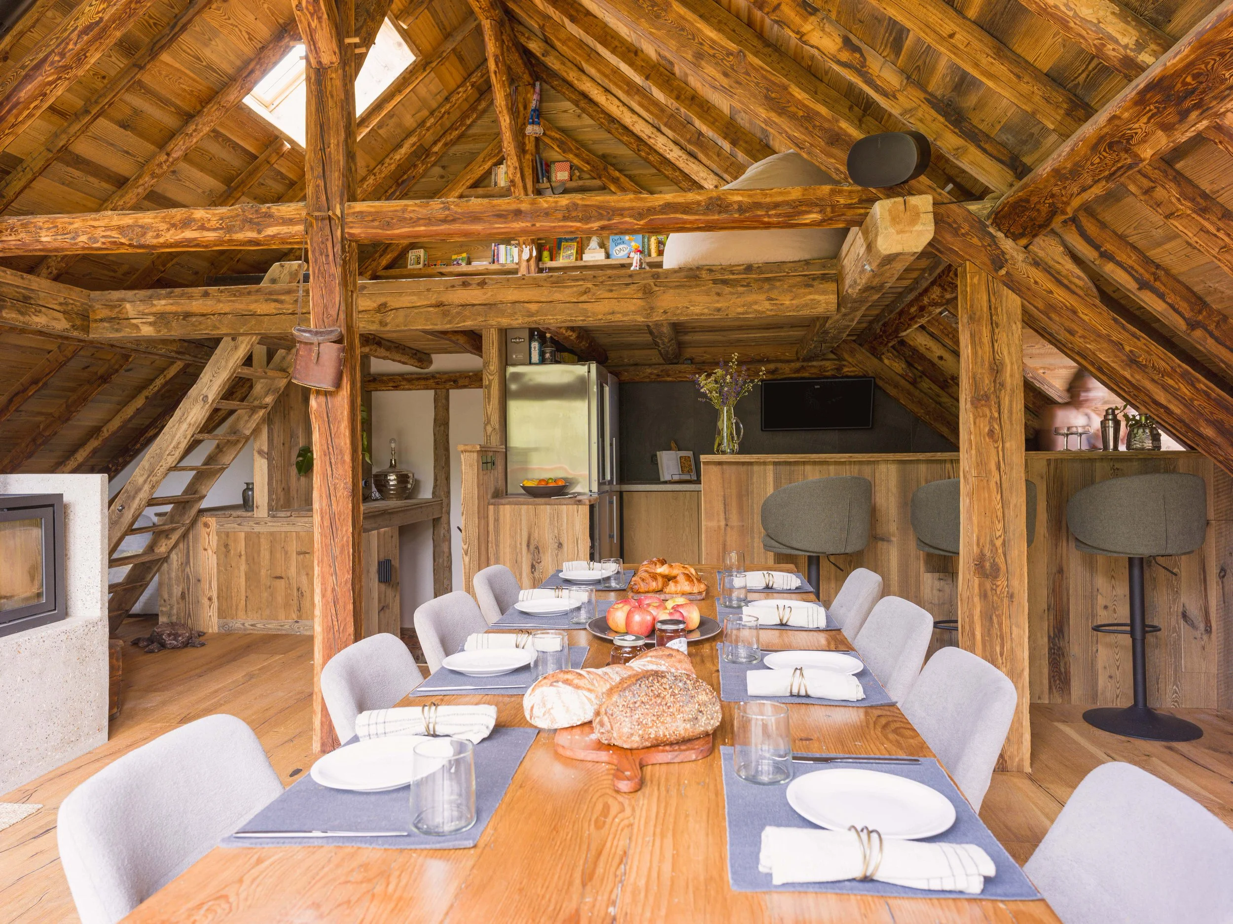 Intérieur d'une cabane rustique en bois avec une table dressée pour le repas, sur laquelle sont disposés du pain, des pommes, des verres et des assiettes ; on aperçoit des poutres apparentes, une mezzanine avec des livres et une cuisine à l'arrière-plan.