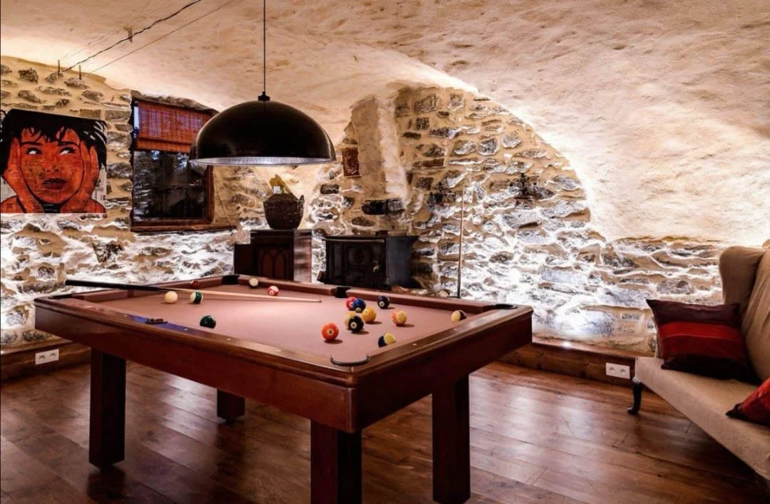 Interior room with a pool table, a painting of a woman's face on the stone wall, a hanging lamp, a window with blinds, a beige couch with striped pillows, and a stone fireplace.