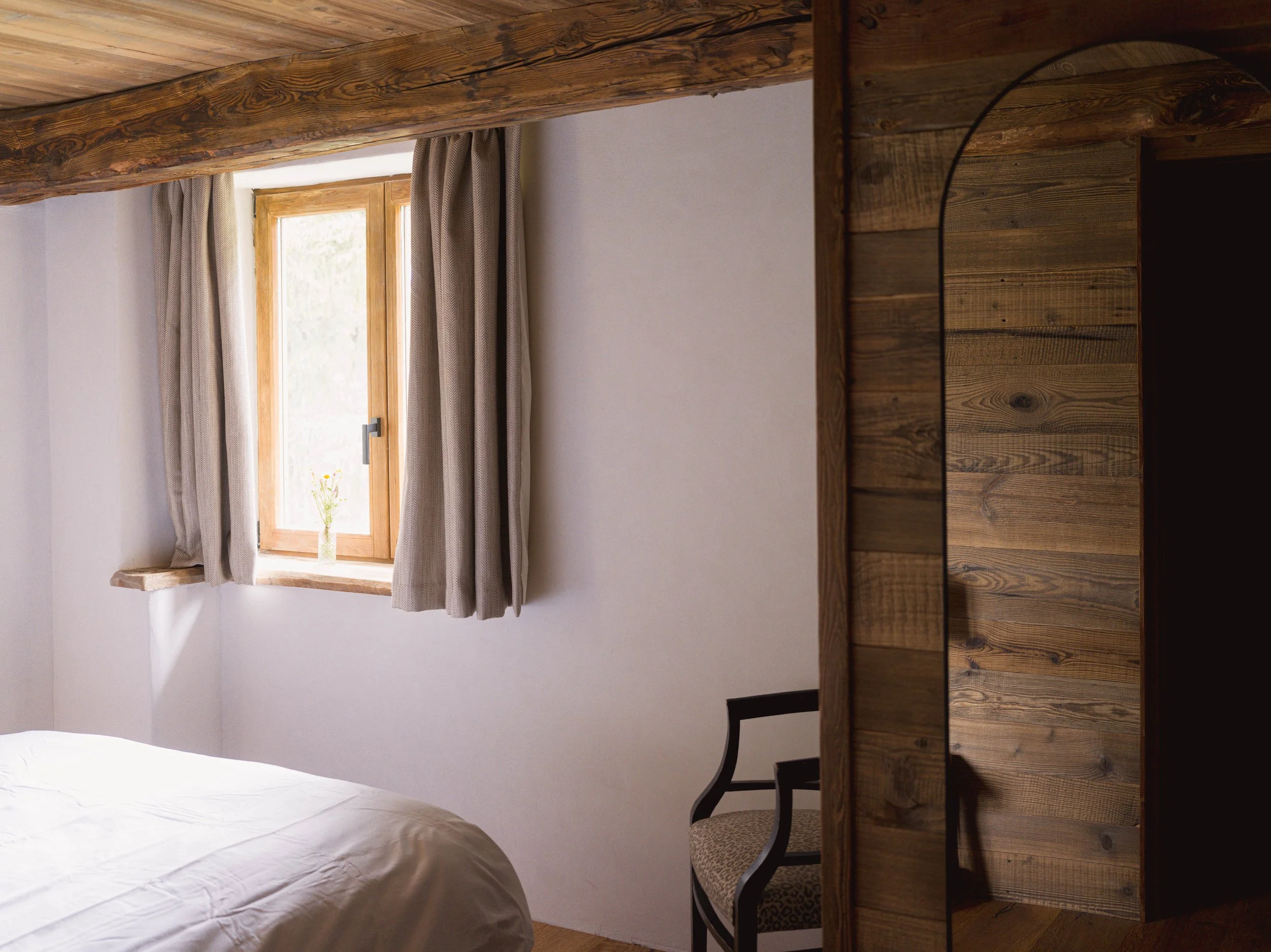 Une chambre avec une poutre apparente et une fenêtre ornée de rideaux, un lit blanc et un bureau en bois avec une chaise.