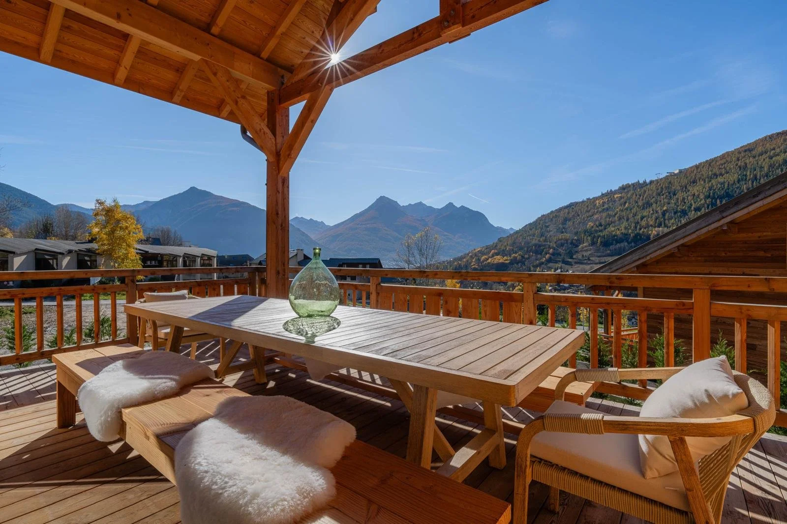 Wooden balcony with a dining table, chairs, and benches overlooking mountains and trees on a sunny day.