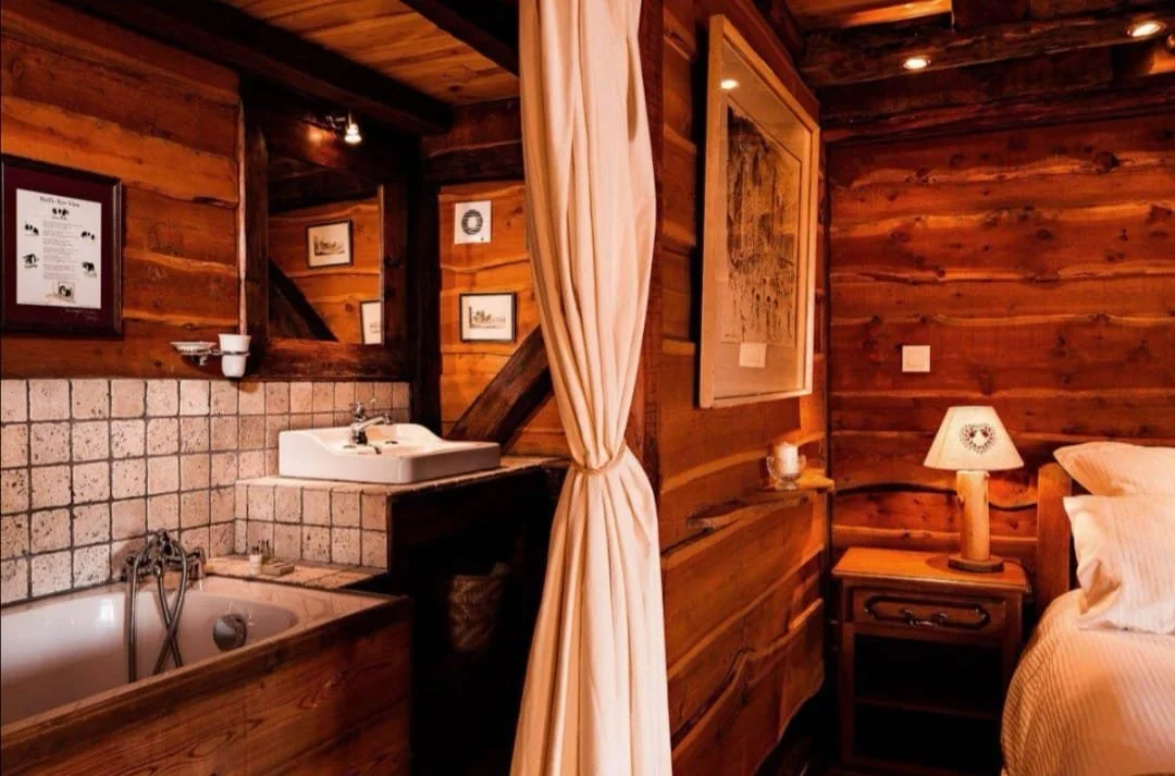 Cozy rustic bedroom with wooden walls, a bedside table with a lamp, a bed with white linens, and a curtain dividing the bed from a small bathroom area that has a sink and bathtub with tiled surround.