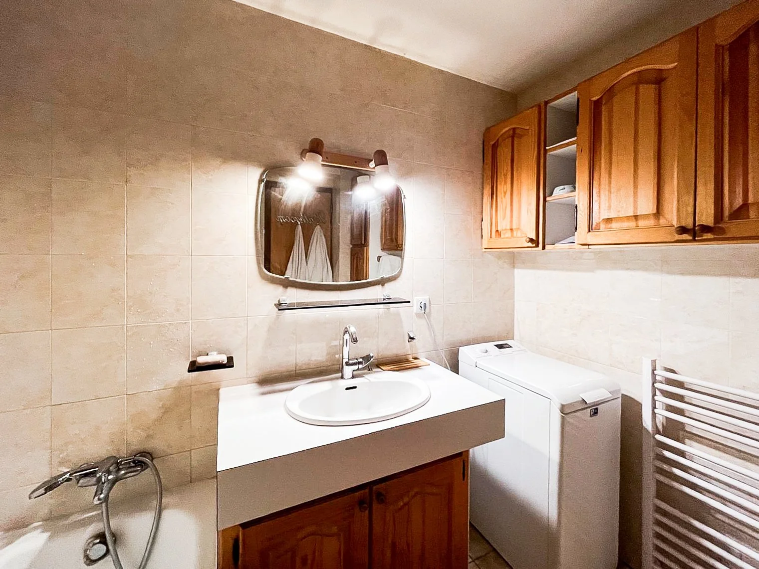 A bathroom featuring a white vanity with a sink, a mirror with lights above, wooden cabinets, a washing machine, and a heated towel rack.