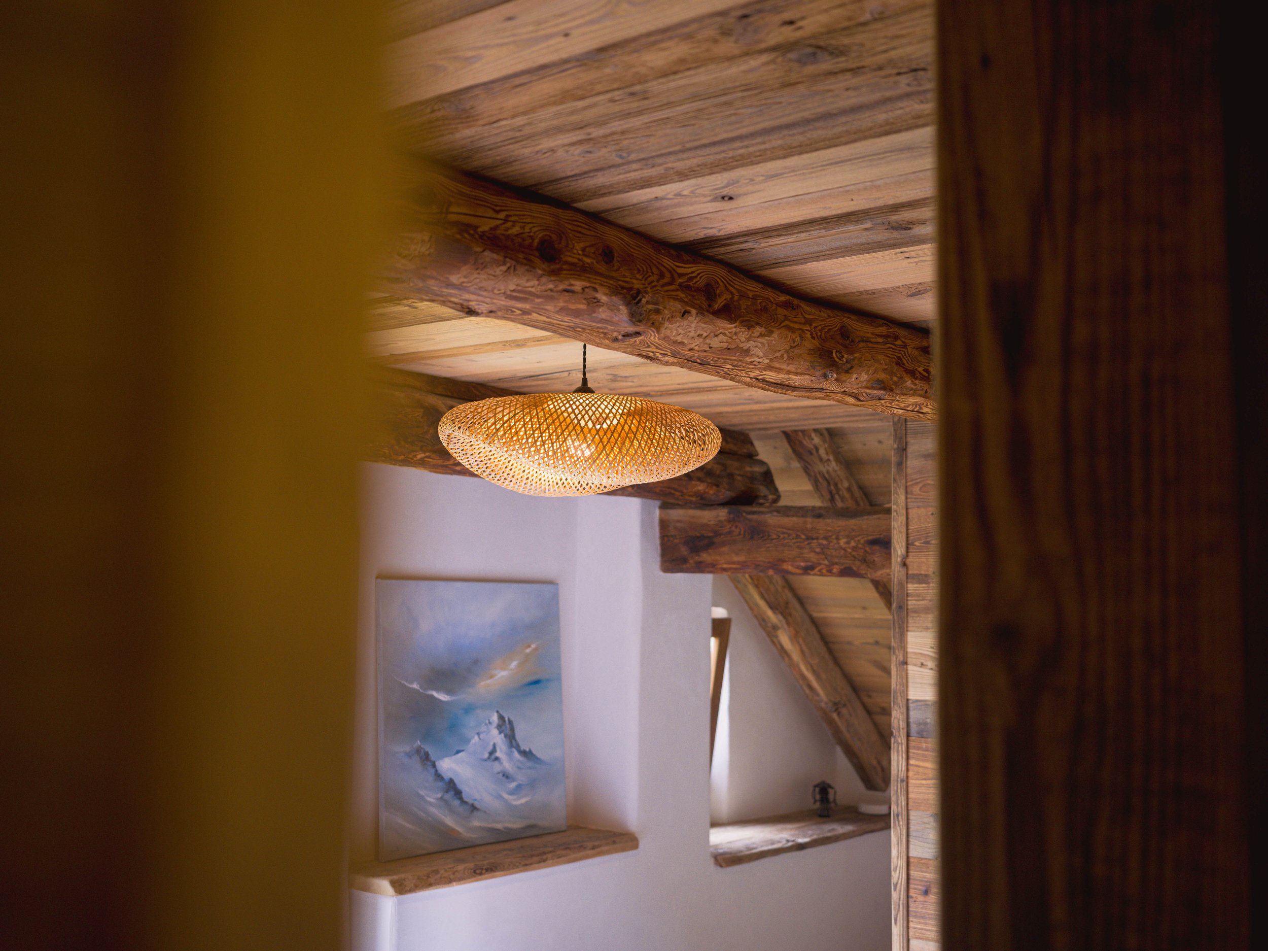 Vue d'un plafond en bois avec un abat-jour tissé suspendu et un tableau représentant un paysage de montagnes enneigées.