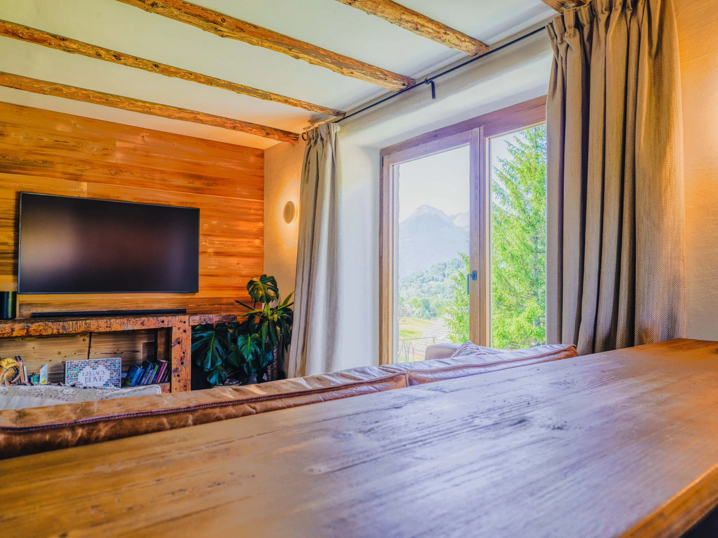 A cozy room featuring a large window with a mountain view, cream curtains, a wooden bed frame, and a flat-screen TV mounted on a wooden-paneled wall. There are houseplants and books underneath the TV, with natural light illuminating the space.