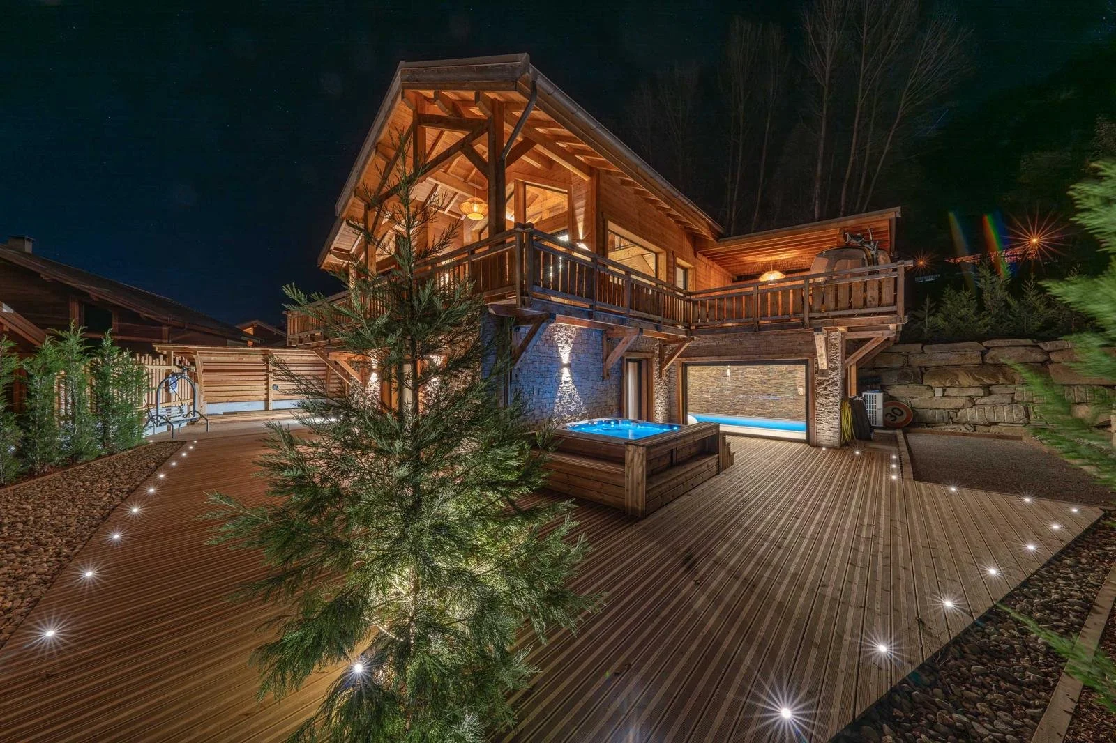 Night view of a wooden two-story house with a deck, illuminated outdoor hot tub, deck lighting, surrounded by trees and rocks.